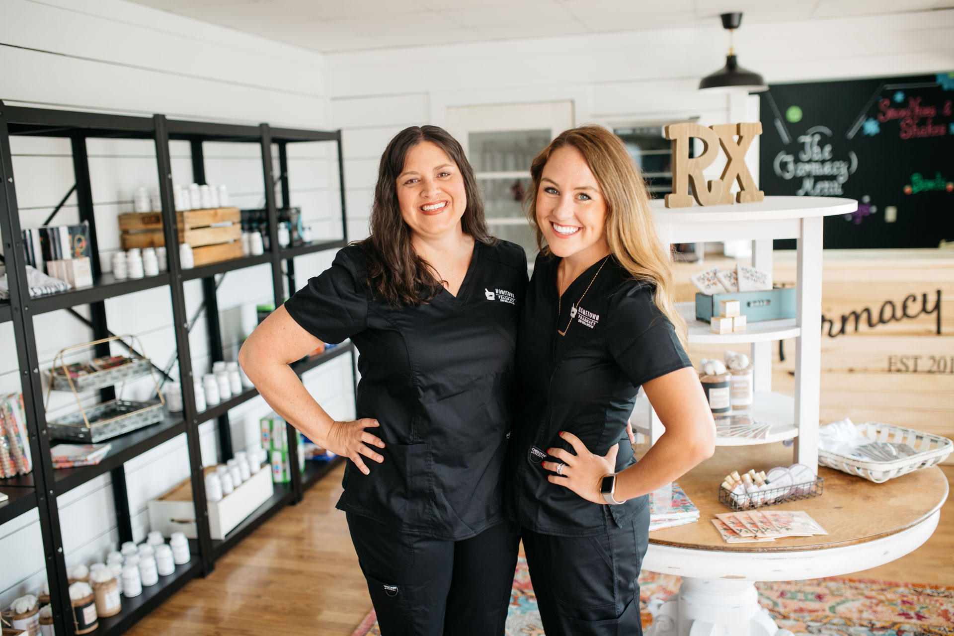 Two smiling Hometown Health System staff members wearing branded black scrubs inside a modern pharmacy store.