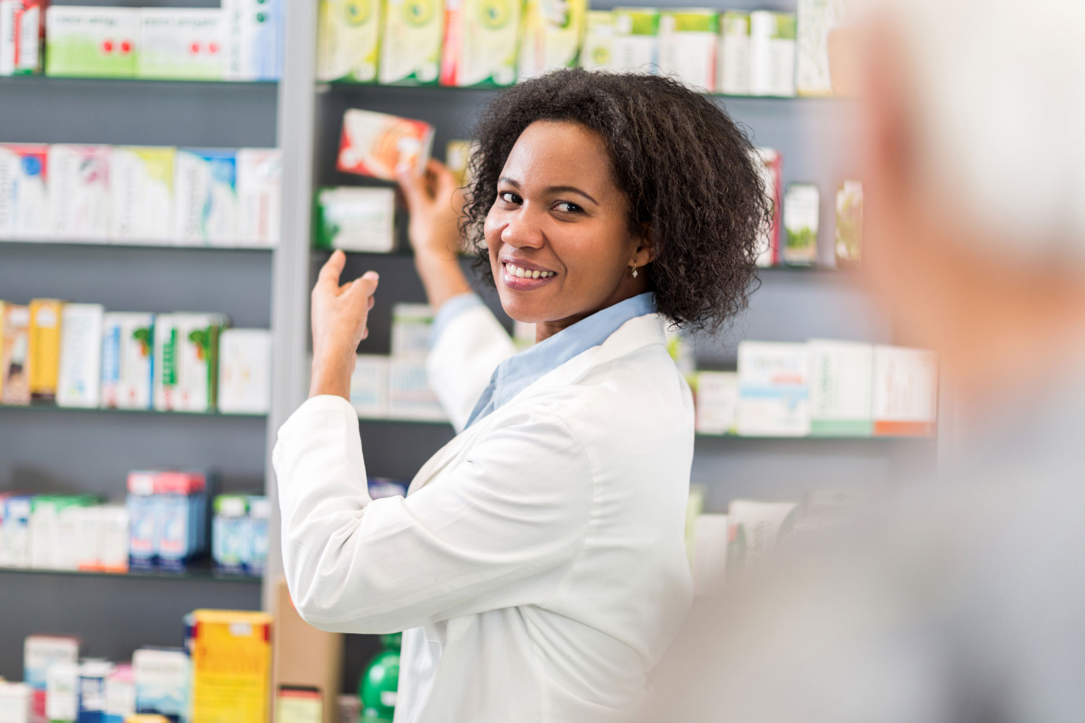 A smiling Black female pharmacist reaching for medication on pharmacy shelves, representing Hometown Health System's specialty pharmacy services.