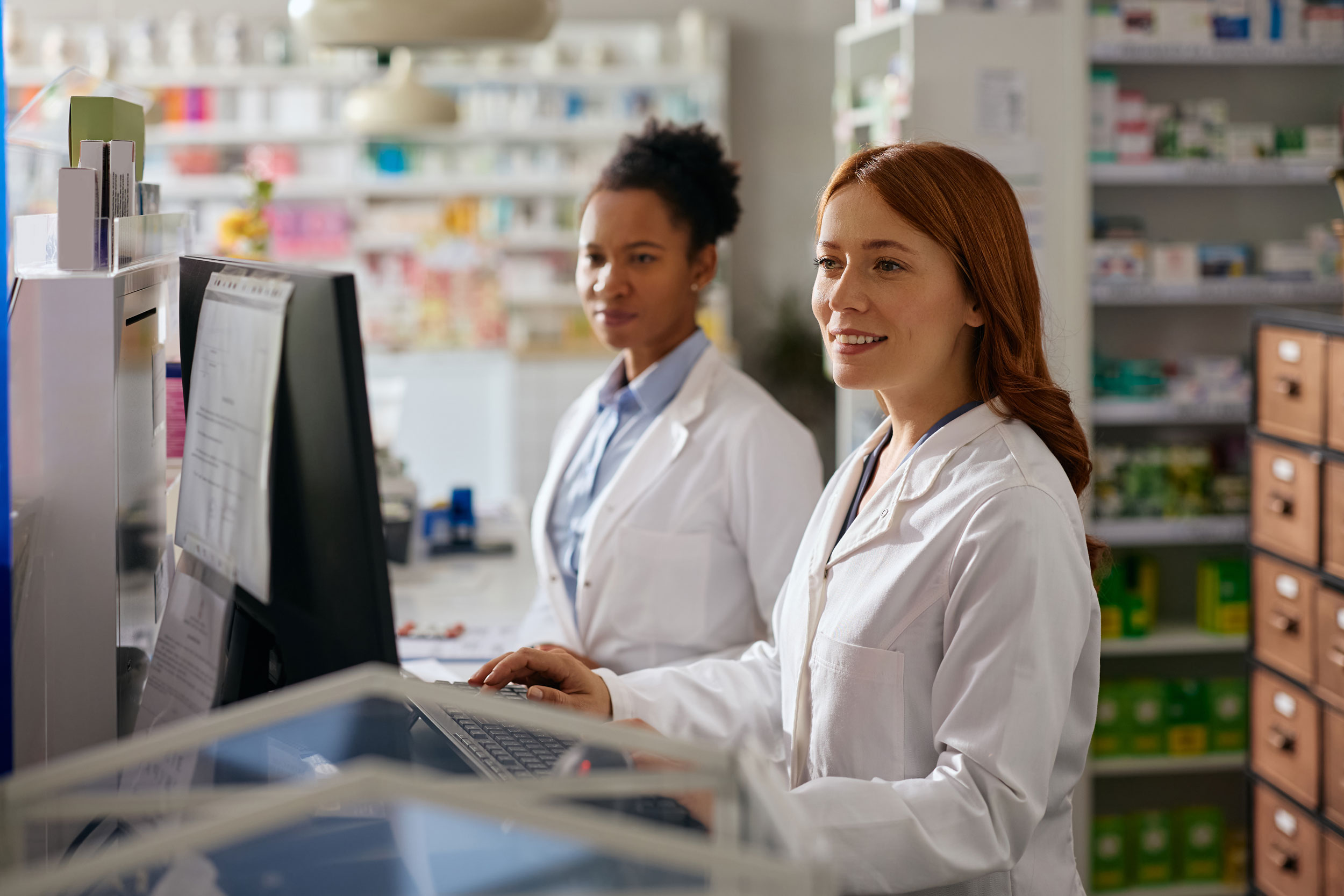 Two female healthcare professionals working at computer stations in a pharmacy, illustrating Hometown Health System's connected care services.