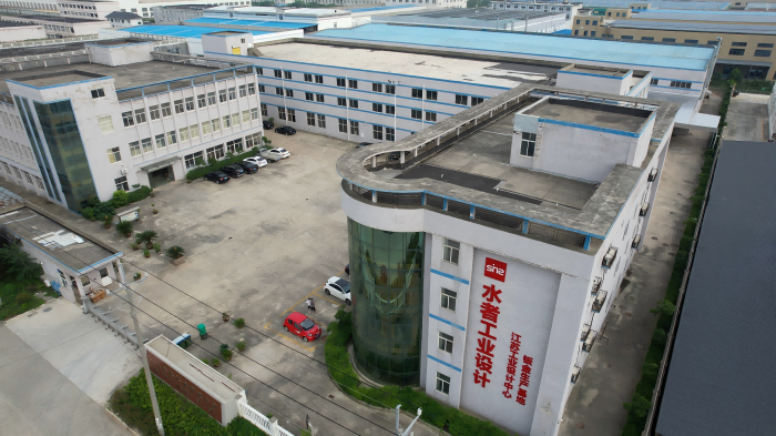 Aerial view of a large industrial building complex with blue roofs and a courtyard with parked cars.