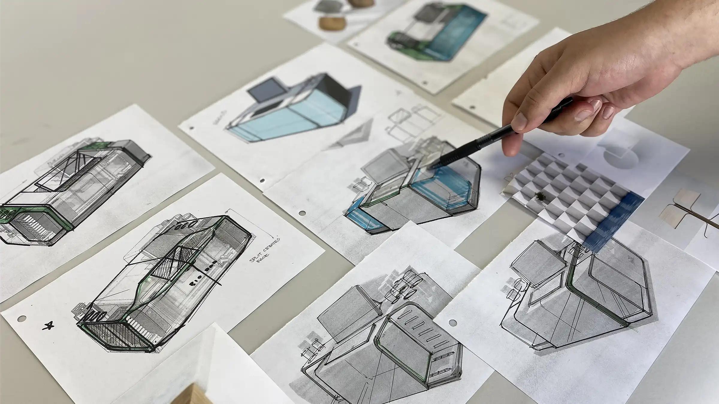 Hand pointing with pen at sketches of product designs with blue and green accents on paper sheets.