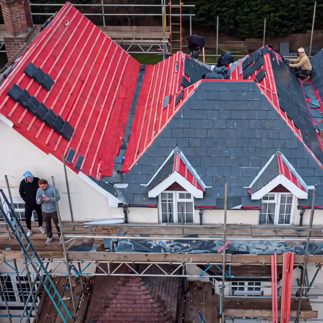 roof replacement in progress with workers installing battens and slate tiles on pitched residential roof with scaffolding and underlayment exposed