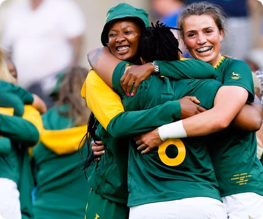 Three female rugby players joyfully hugging, wearing South African green and yellow jerseys.