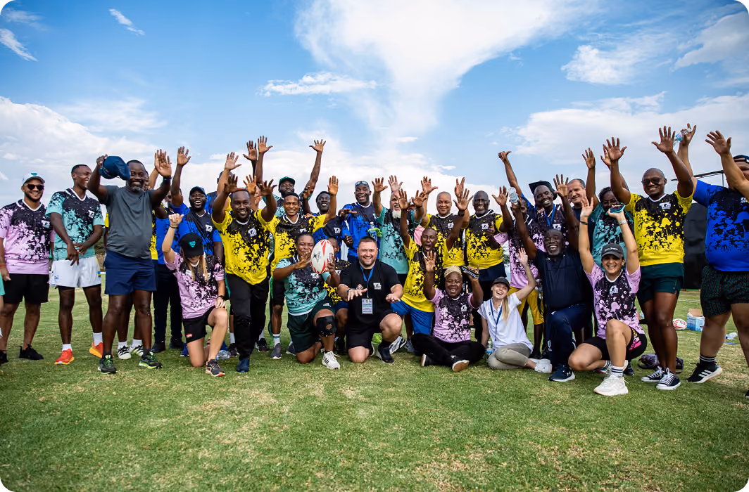 Large group of smiling people in colorful sportswear raising hands on a grassy field under blue sky.