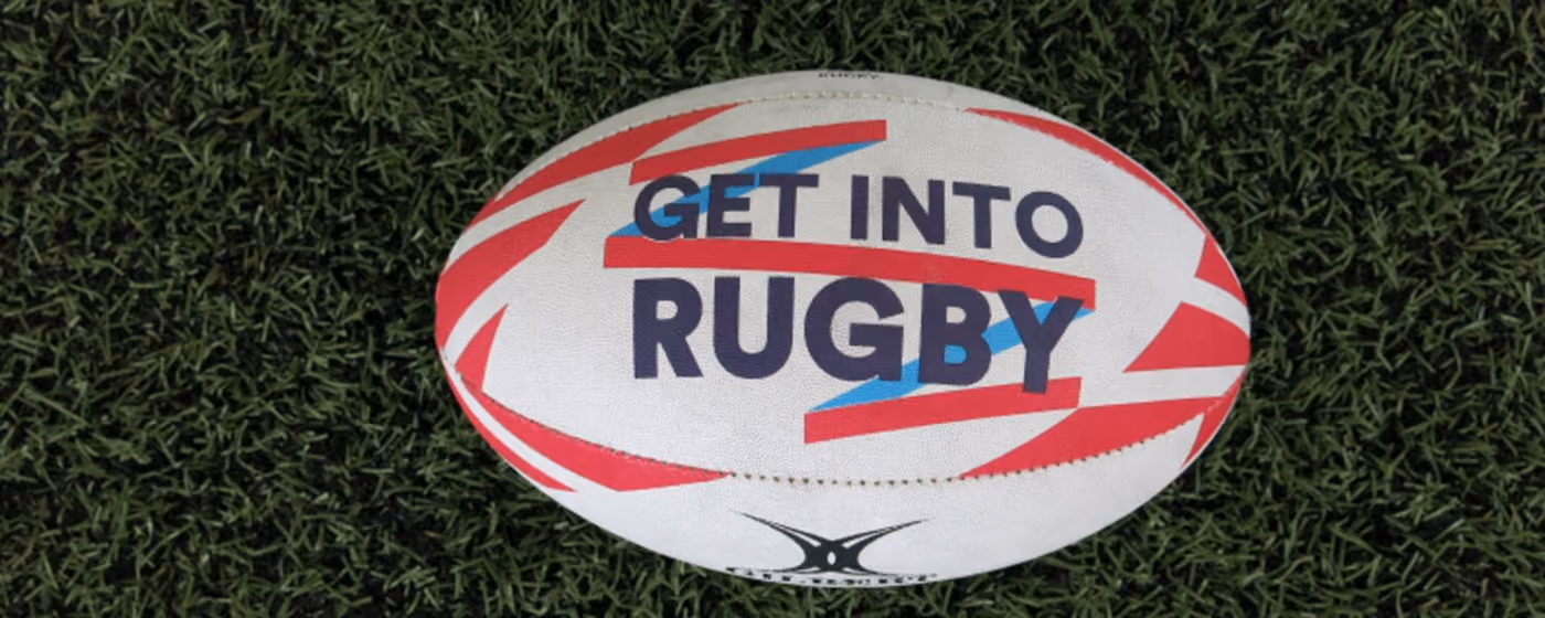 White rugby ball with red and blue accents and text 'Get into Rugby' on green grass.