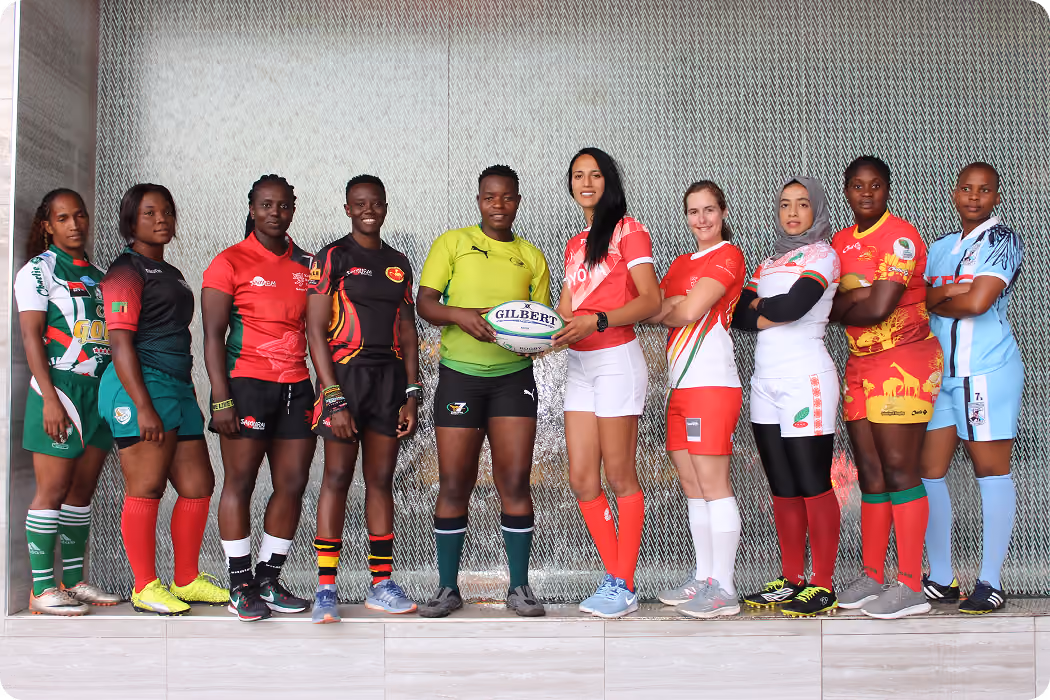 Ten women rugby players in colorful uniforms standing side by side, one holding a rugby ball.