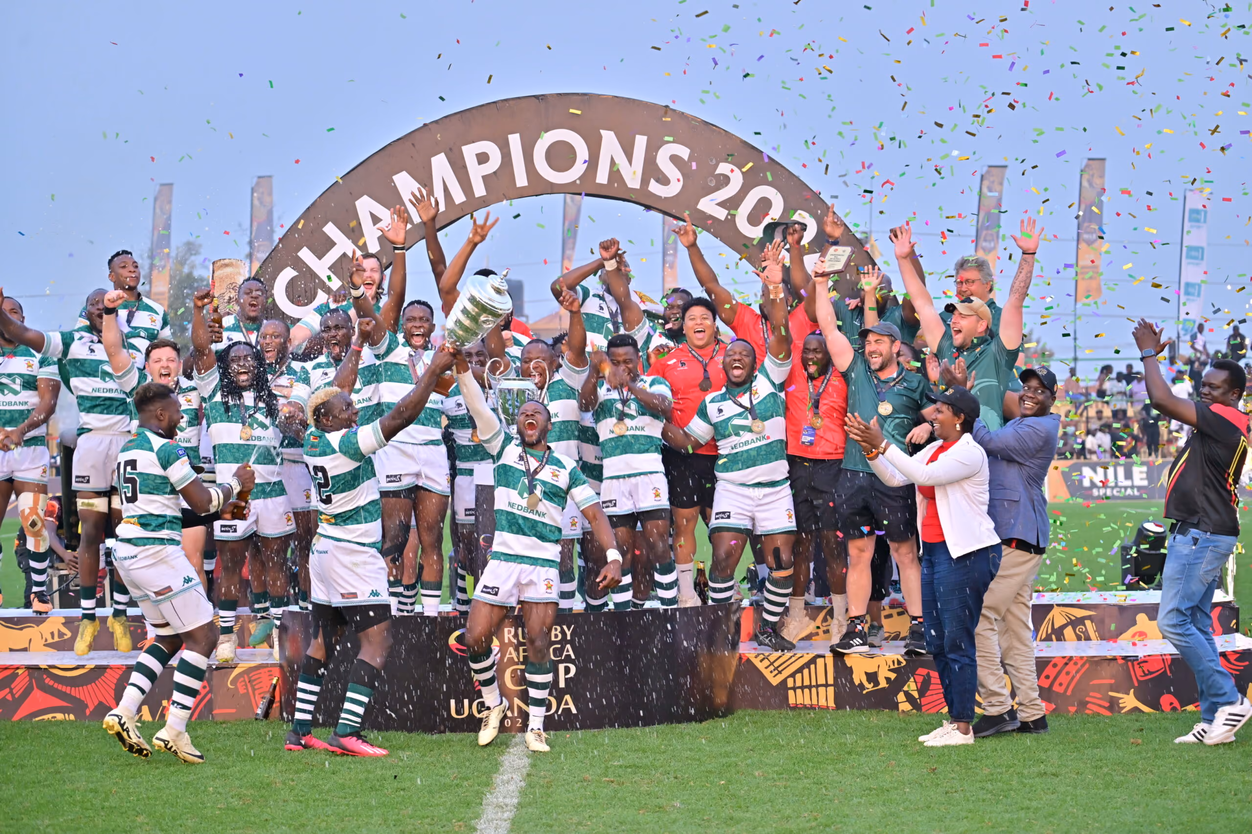 Rugby team celebrating their victory as champions 2023, holding up a trophy on a grassy field under a sign that reads CHAMPIONS 2023, with confetti falling around them.