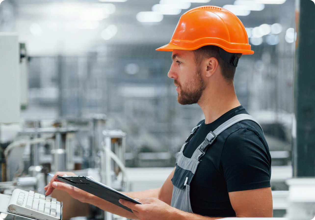 Man in an orange hard hat and gray overalls using a keyboard and holding a clipboard in a factory.
