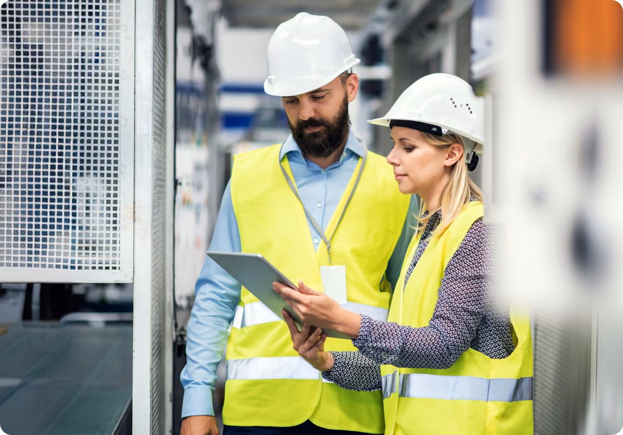 Two factory workers in safety vests and helmets reviewing a tablet in an industrial setting.