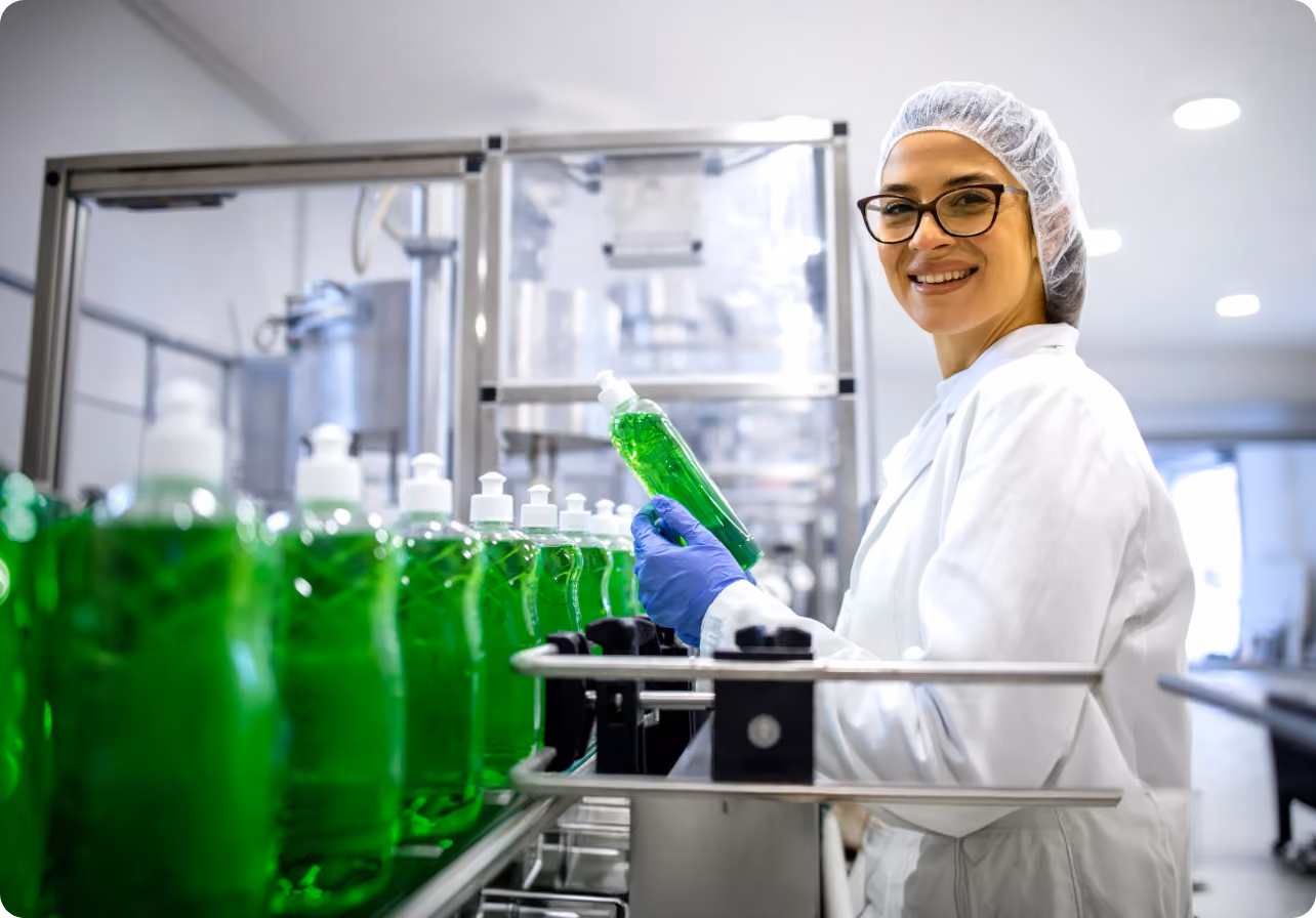 Smiling factory worker in protective gear inspecting green liquid soap bottles on production line.