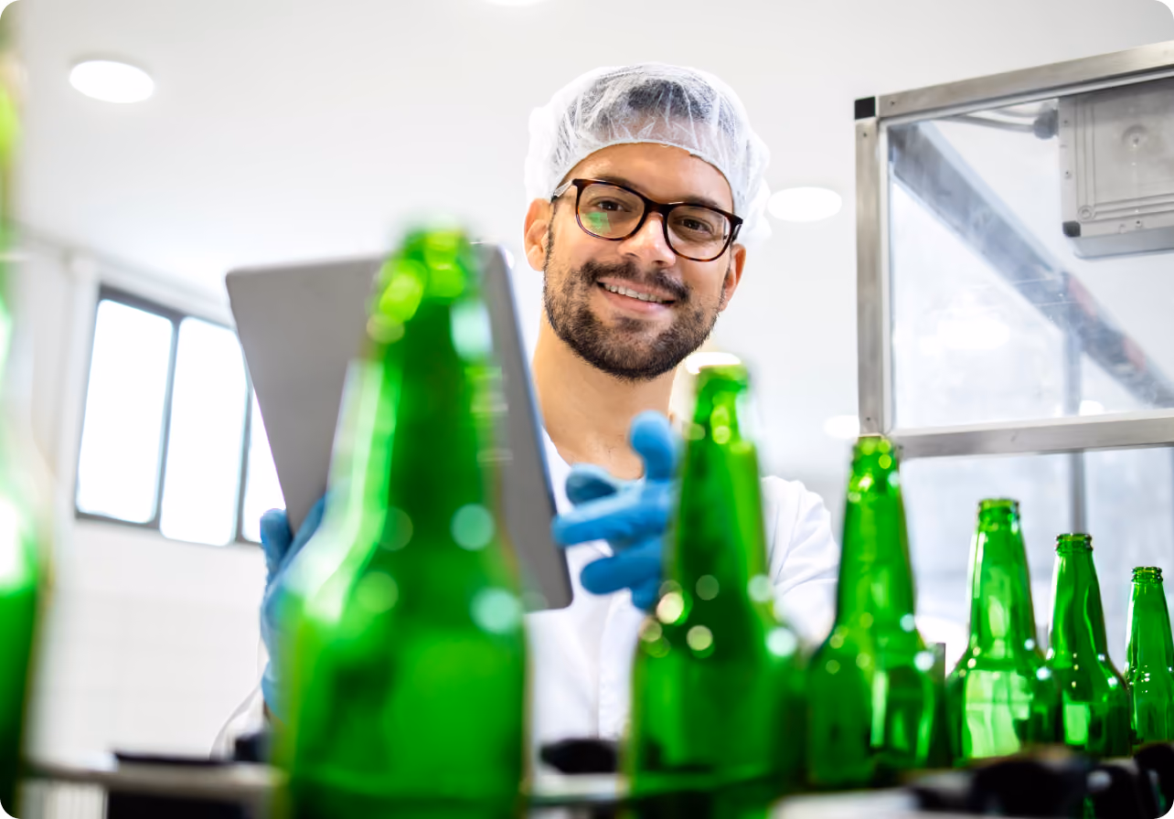 Man in hairnet and gloves uses tablet to inspect green glass bottles in a clean production facility.