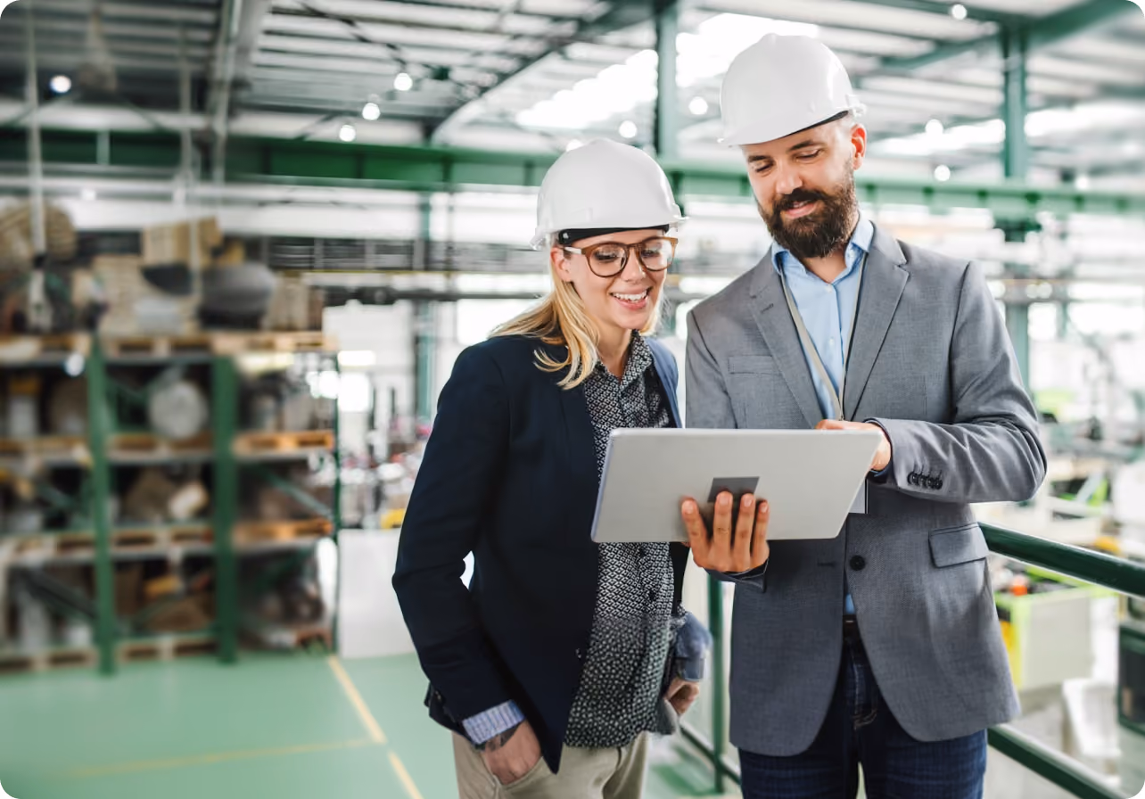 Two supervisors in hard hats looking at a tablet inside a warehouse or industrial facility.