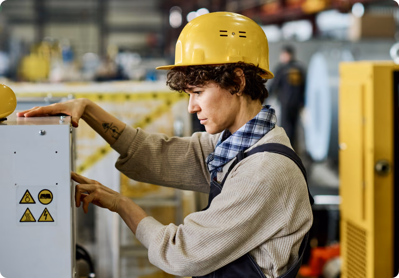 Worker in a yellow hard hat adjusting controls on industrial machinery in a factory.
