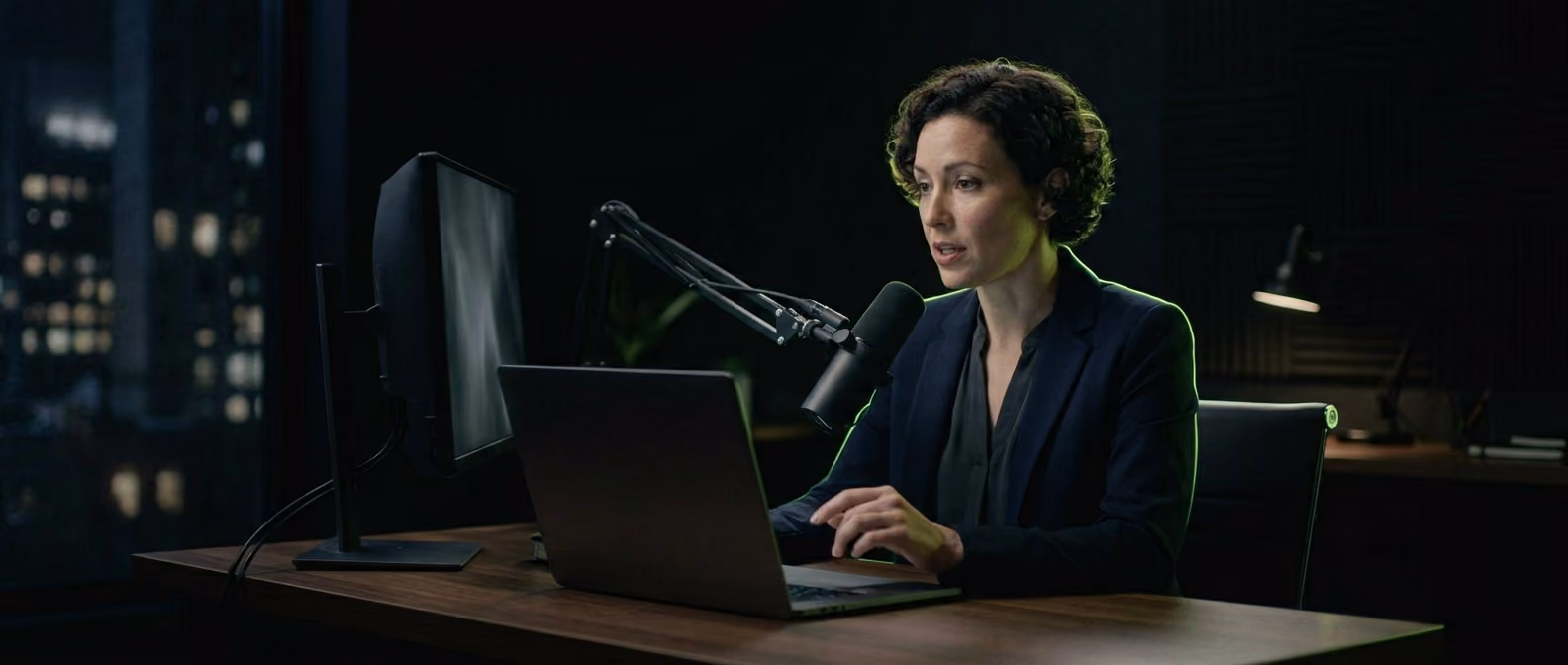 A presenter sits in a dark home-studio setup, face lit by the glow of a laptop mid-webinar, soft lime-green rim light on the desk edge.