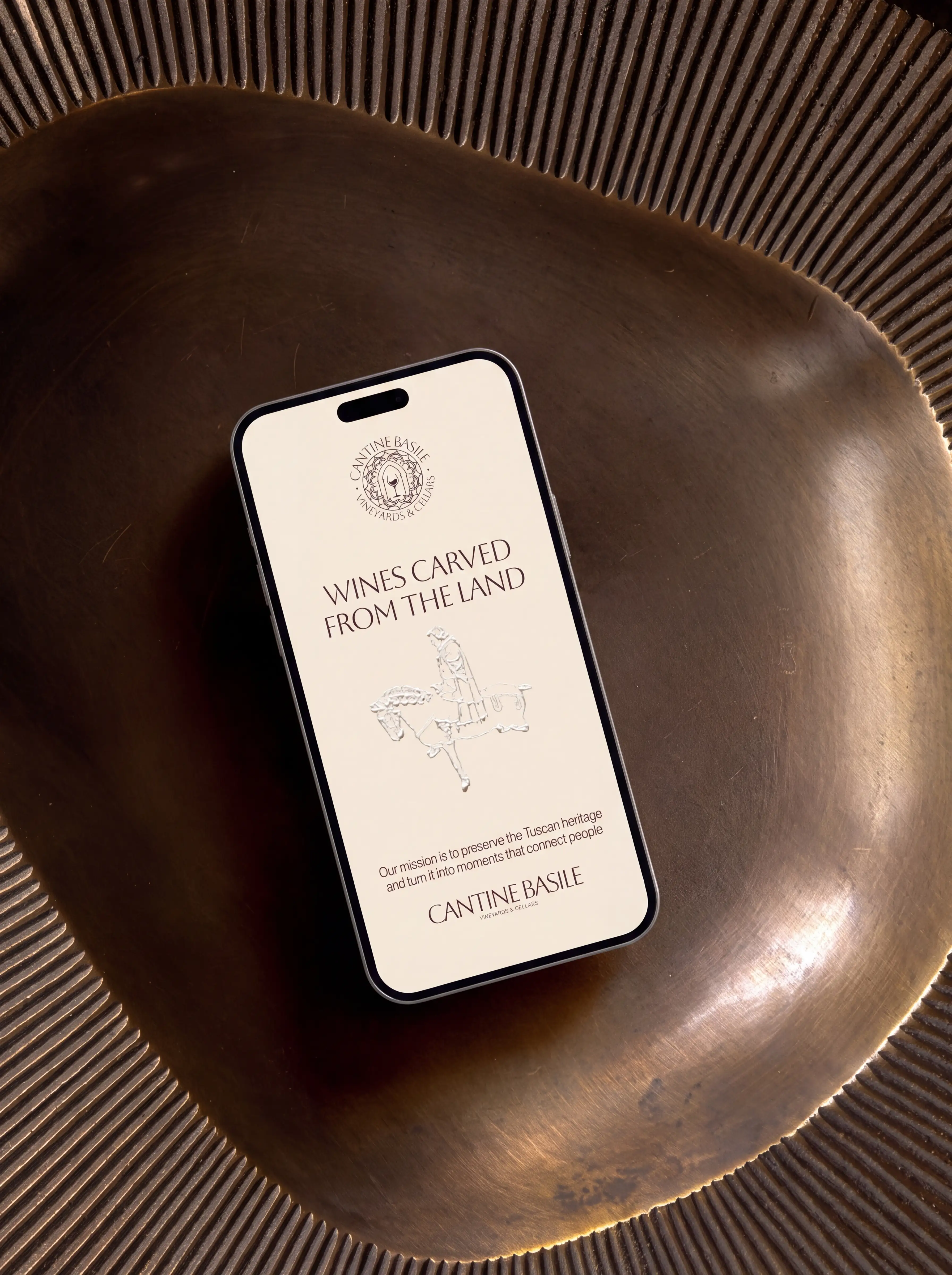 Smartphone screen displaying Cantine Basile Vineyards & Cellars branding with the text 'WINES CARVED FROM THE LAND' and a mission statement about preserving Tuscan heritage, placed on a textured bronze metal surface.