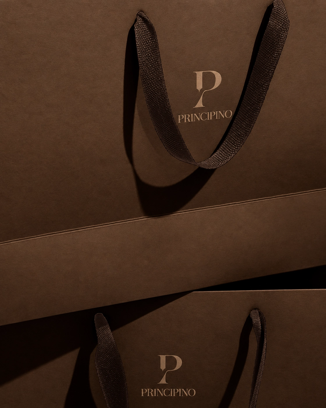 Close-up of two overlapping brown shopping bags with dark textured handles, each bag displaying the gold logo and text 'PRINCIPINO'.