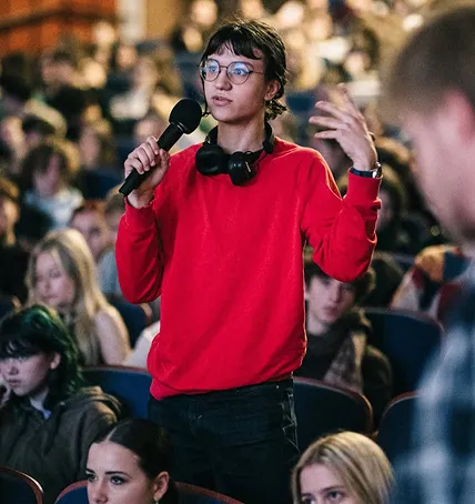 Mladý člověk v červeném svetru mluví do mikrofonu na veřejné akci s publikem.