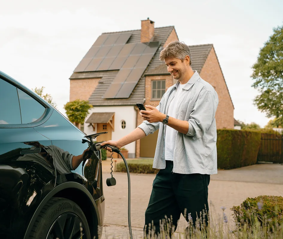 Man plugging in his electric car at home while checking his phone, with solar panels on the roof behind him