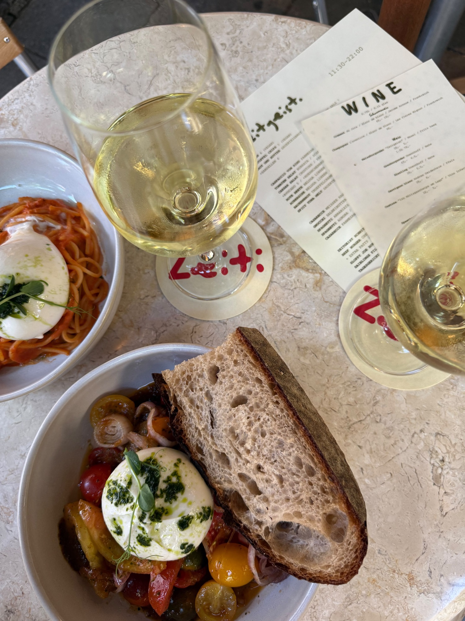 Gedeckter Marmortisch mit Burrata auf Tomaten, Pasta, Sauerteigbrot, zwei Gläsern Weißwein und der Zeitgeist-Weinkarte