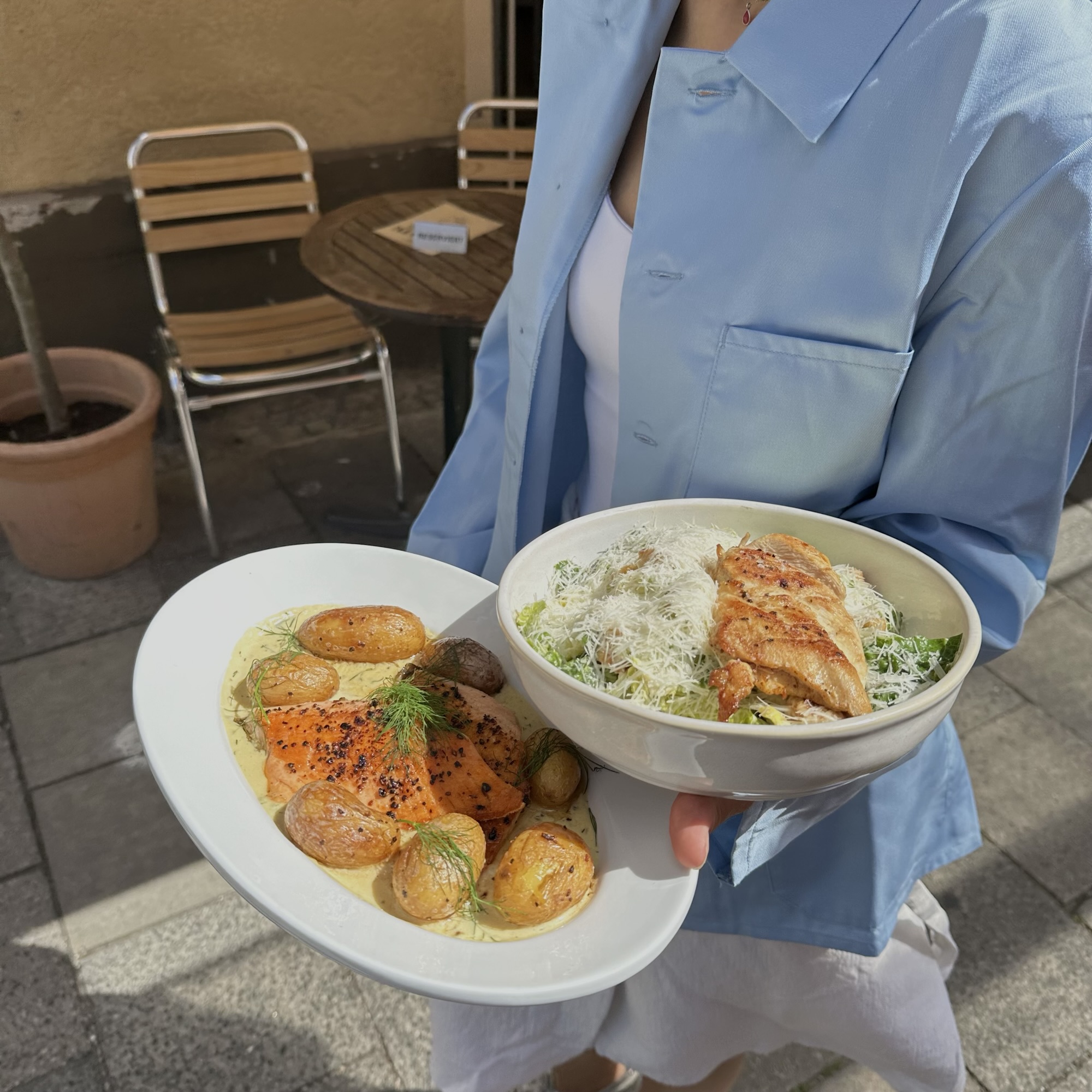 Servicekraft im hellblauen Hemd trägt einen Teller Lachs mit Kartoffeln und eine Caesar-Salat-Bowl zu den Tischen vor dem Lokal