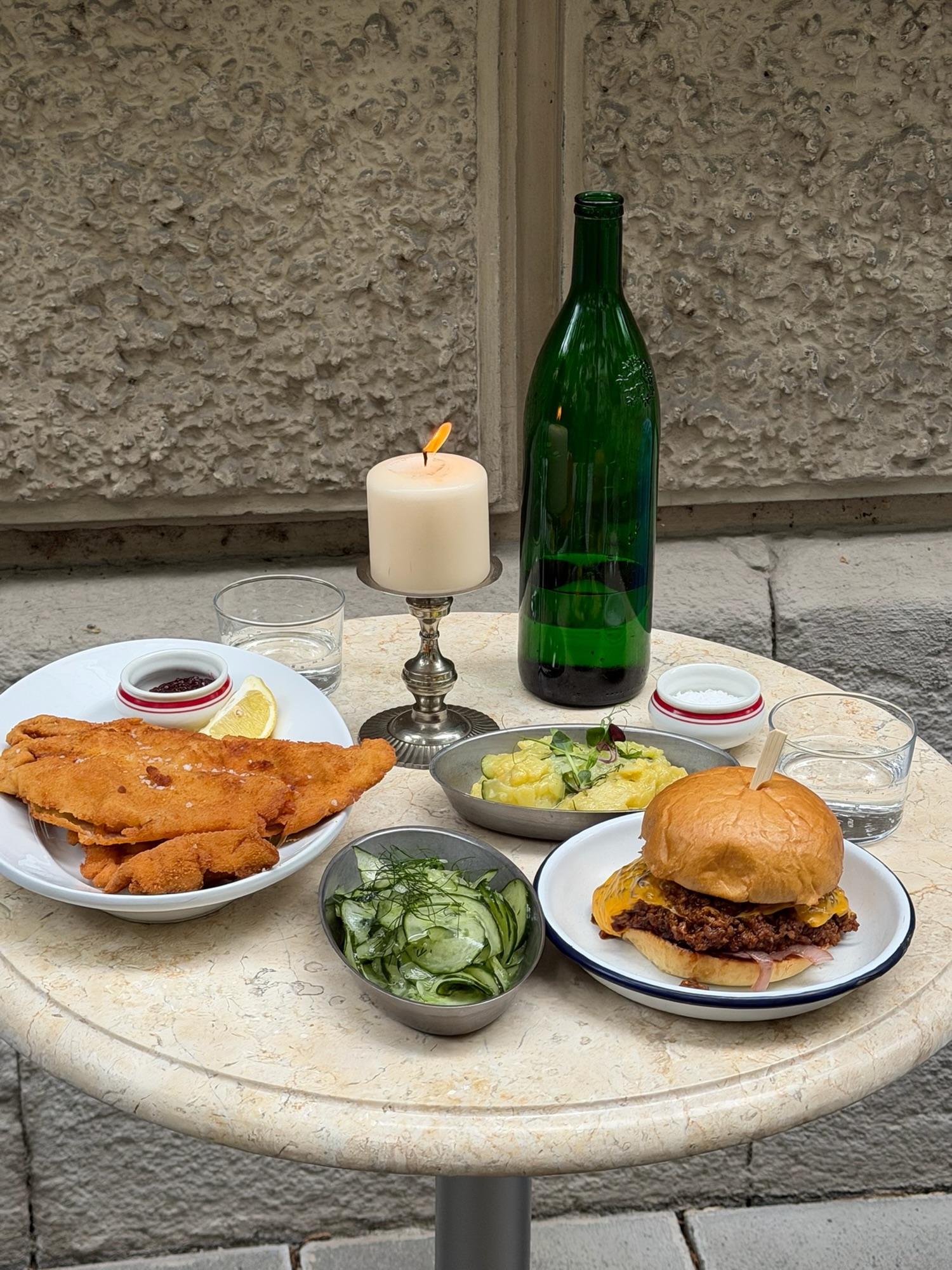 Marmortisch mit Wiener Schnitzel, Kartoffelsalat, Gurkensalat, Sloppy-Joe-Burger, Kerze und grüner Weinflasche