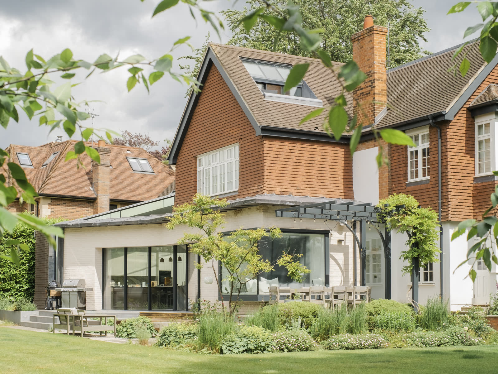 A pale brick single storey extension with wide sliding doors and a steel pergola, set below a tile hung house, with garden planting in the foreground.