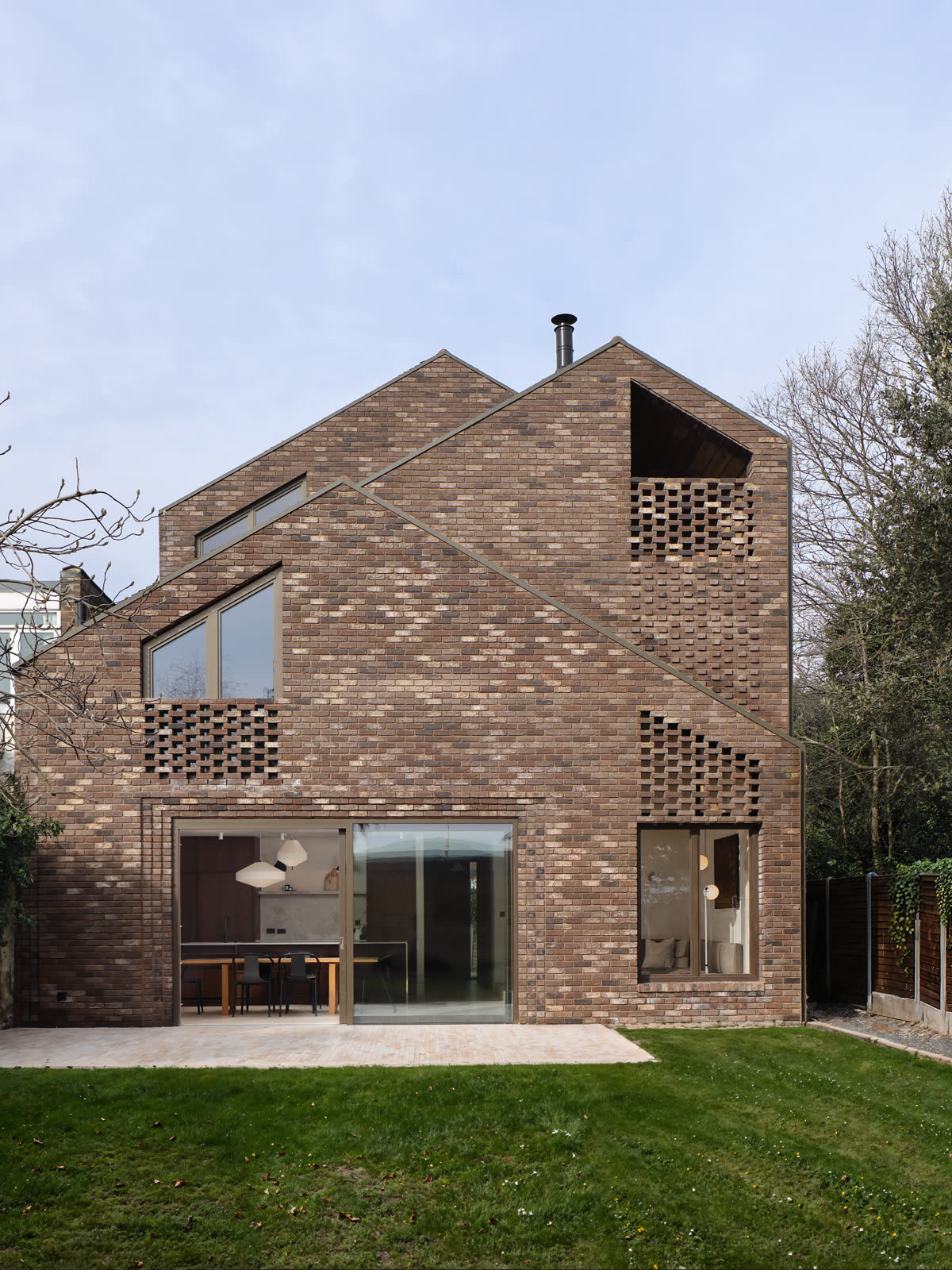 Rear elevation of a brick house with a pitched gable, perforated brick screens and full height sliding doors opening onto a paved terrace and lawn.