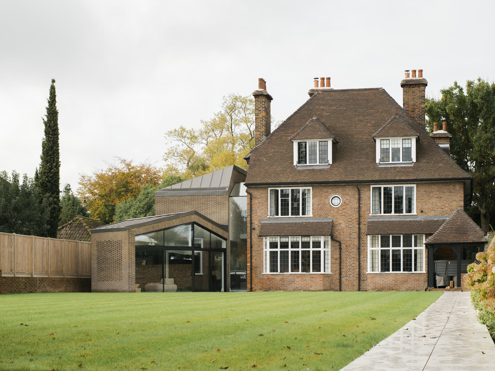 A brick and glass extension with a standing seam roof joining a red brick house with a tiled roof and tall chimneys, seen across the lawn.