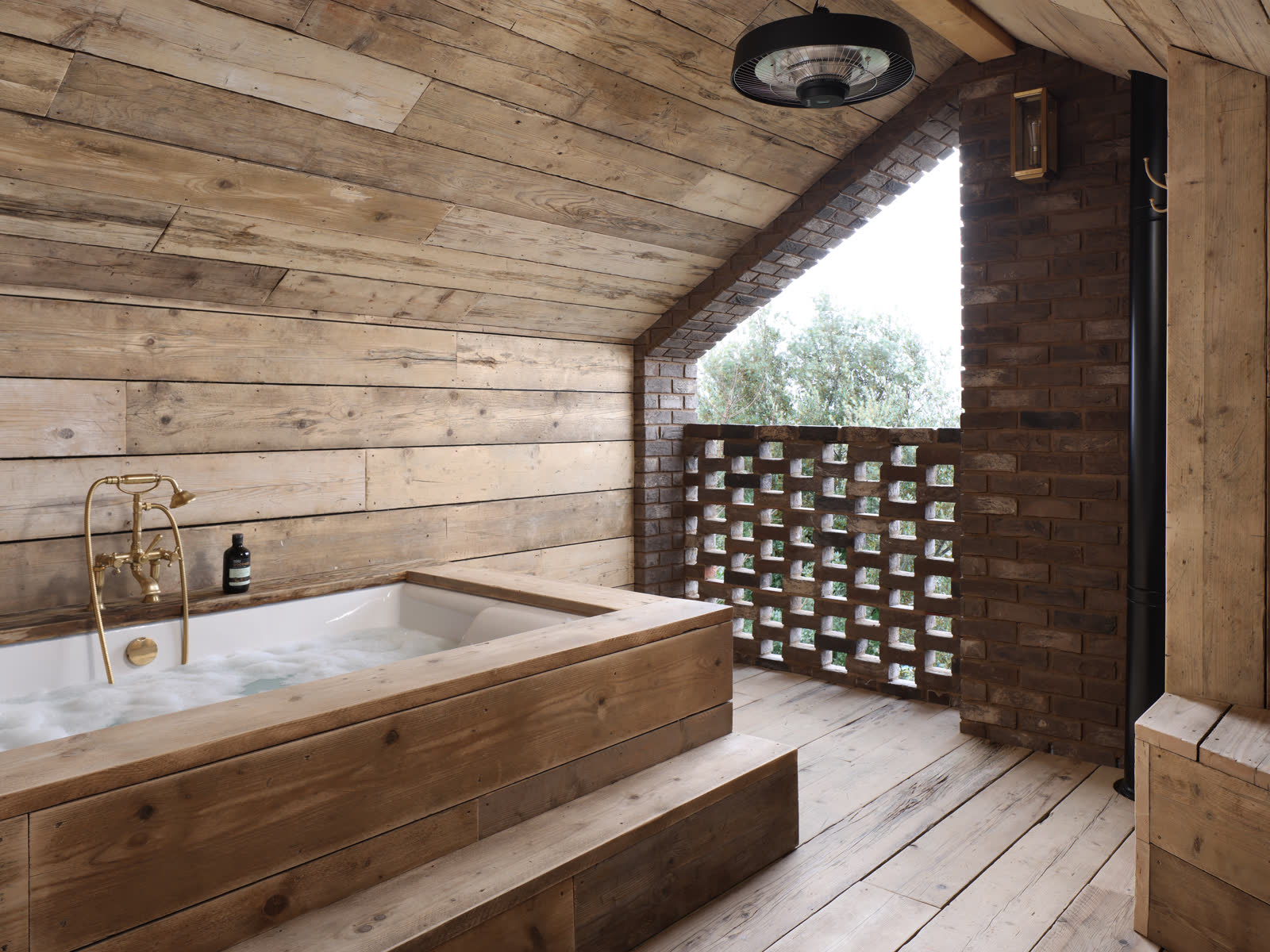 A timber lined loft bathroom with a sunken bath, a brass wall mounted mixer and hand shower, and a perforated brick screen looking onto trees.