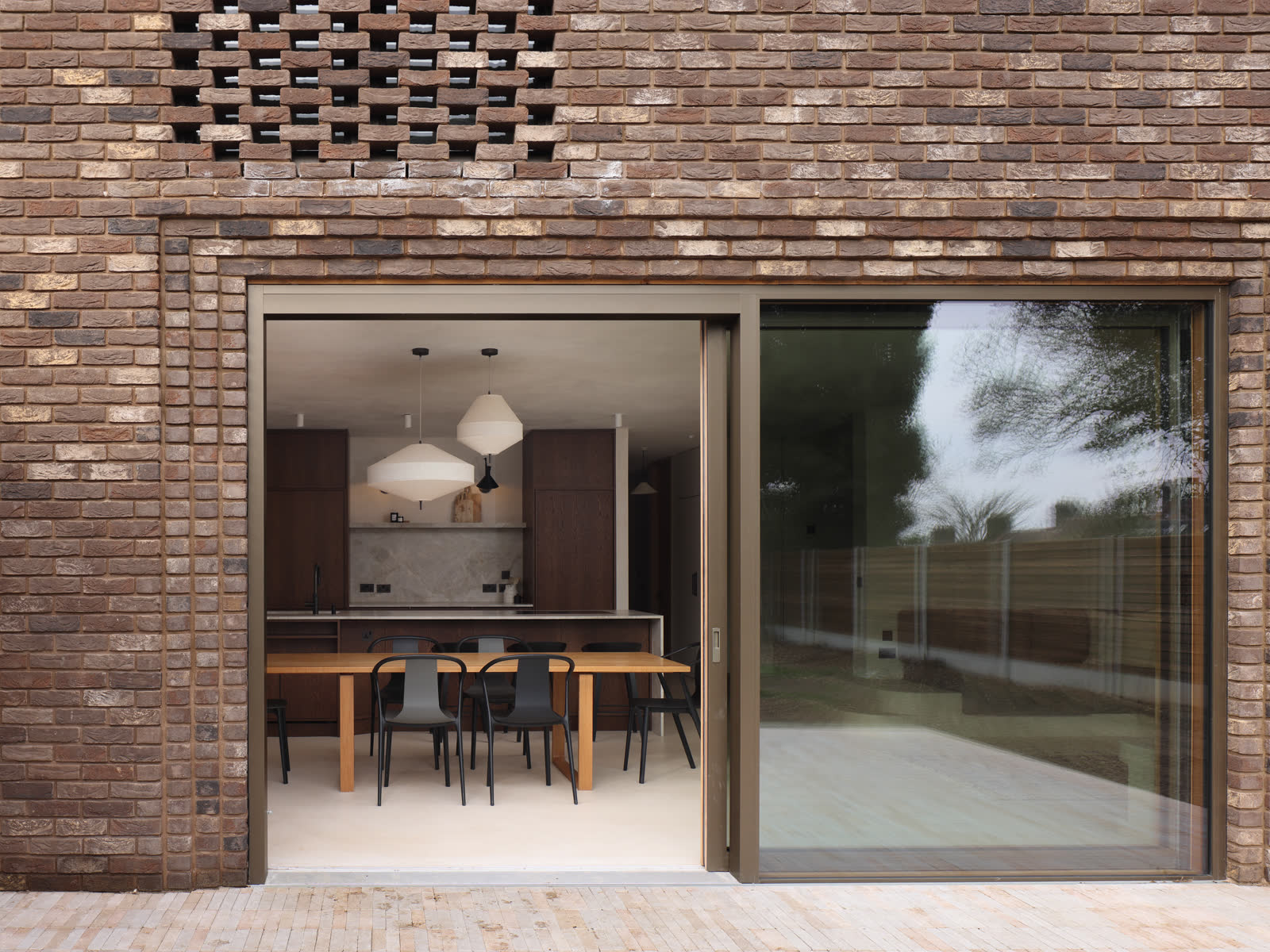 A brick elevation with a perforated brick panel above a wide sliding door, open onto a kitchen and dining room with a timber table and pendant lights.
