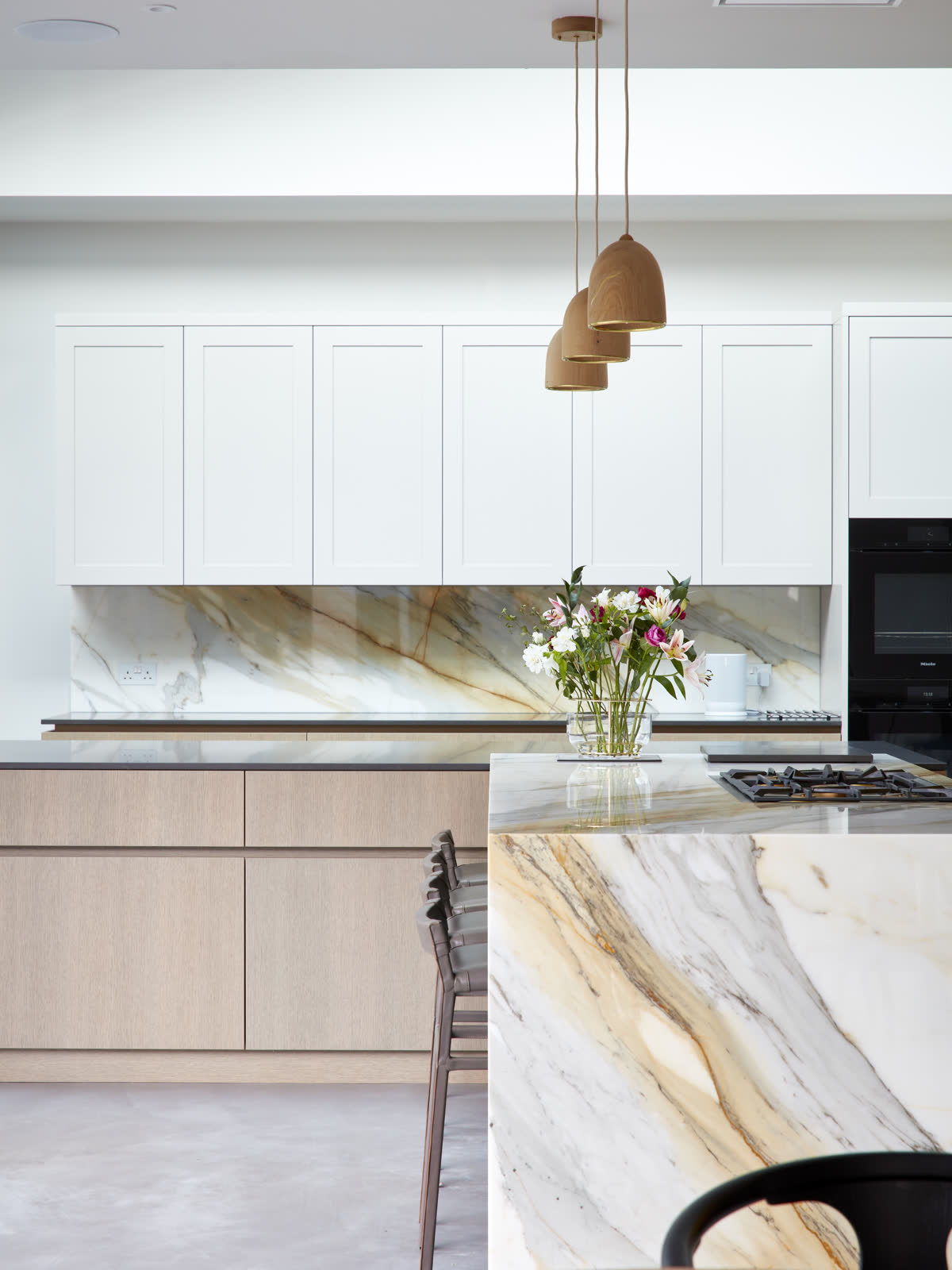 A kitchen with a veined marble splashback and waterfall island, pale oak cabinetry, white wall units and turned wooden pendant lights.