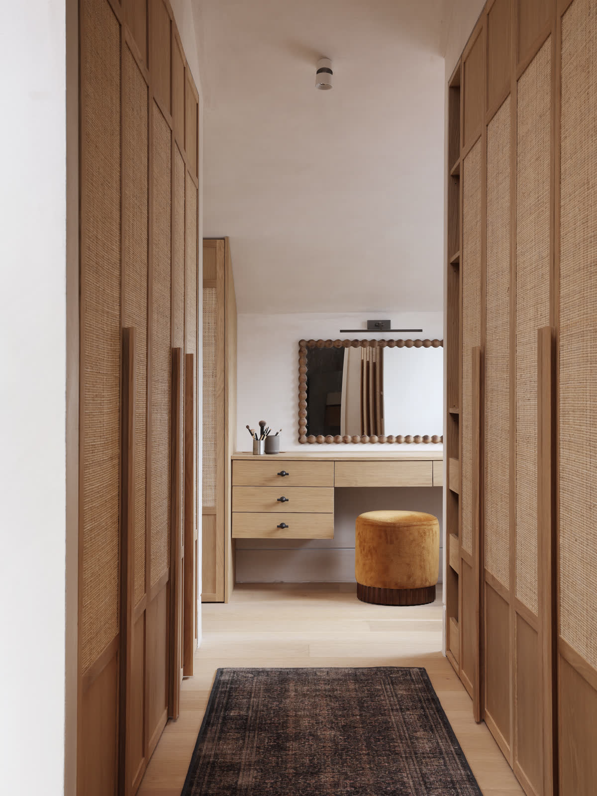 A dressing room lined with oak and rattan wardrobe doors, leading to a dressing table with a bobbin frame mirror and an ochre velvet stool.