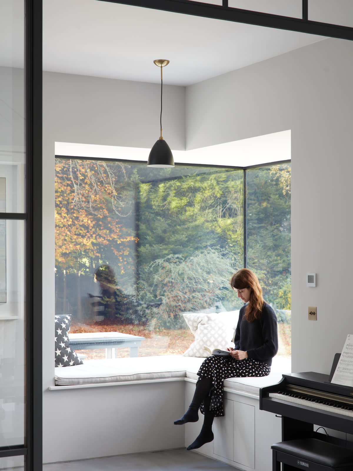 A cushioned window seat in a glazed corner looking onto autumn trees, with a person sitting reading and a piano alongside.