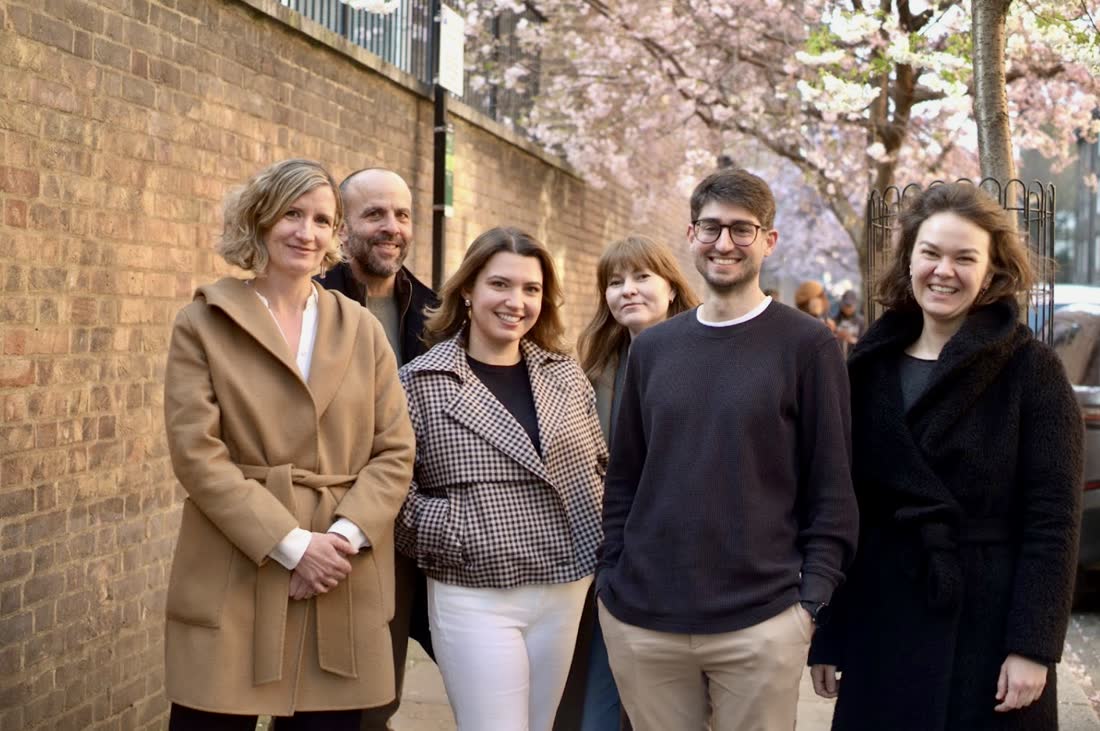 Six members of the team standing together on the pavement beside a brick wall, under a cherry tree in blossom.