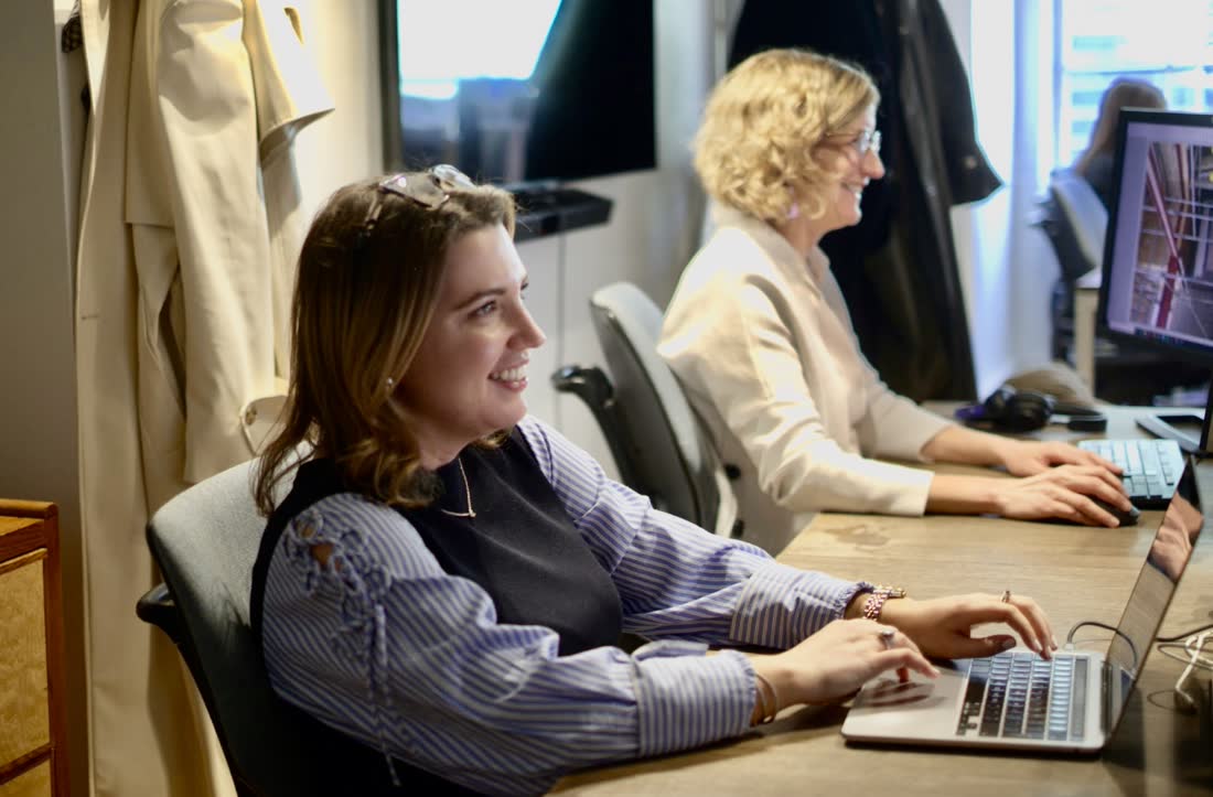 Two colleagues working side by side at a shared desk, one at a laptop and one at a monitor showing a project photograph.