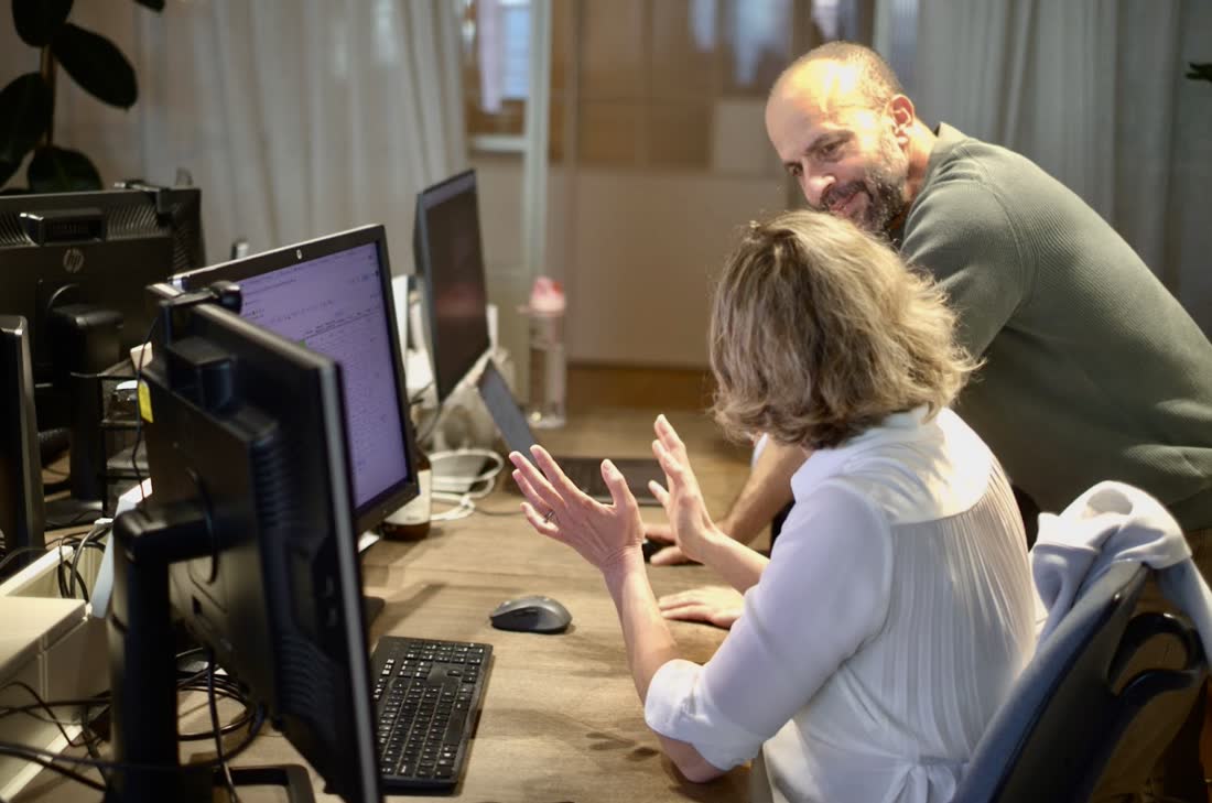 A colleague leaning over a desk to talk something through with a team member seated at their monitors.