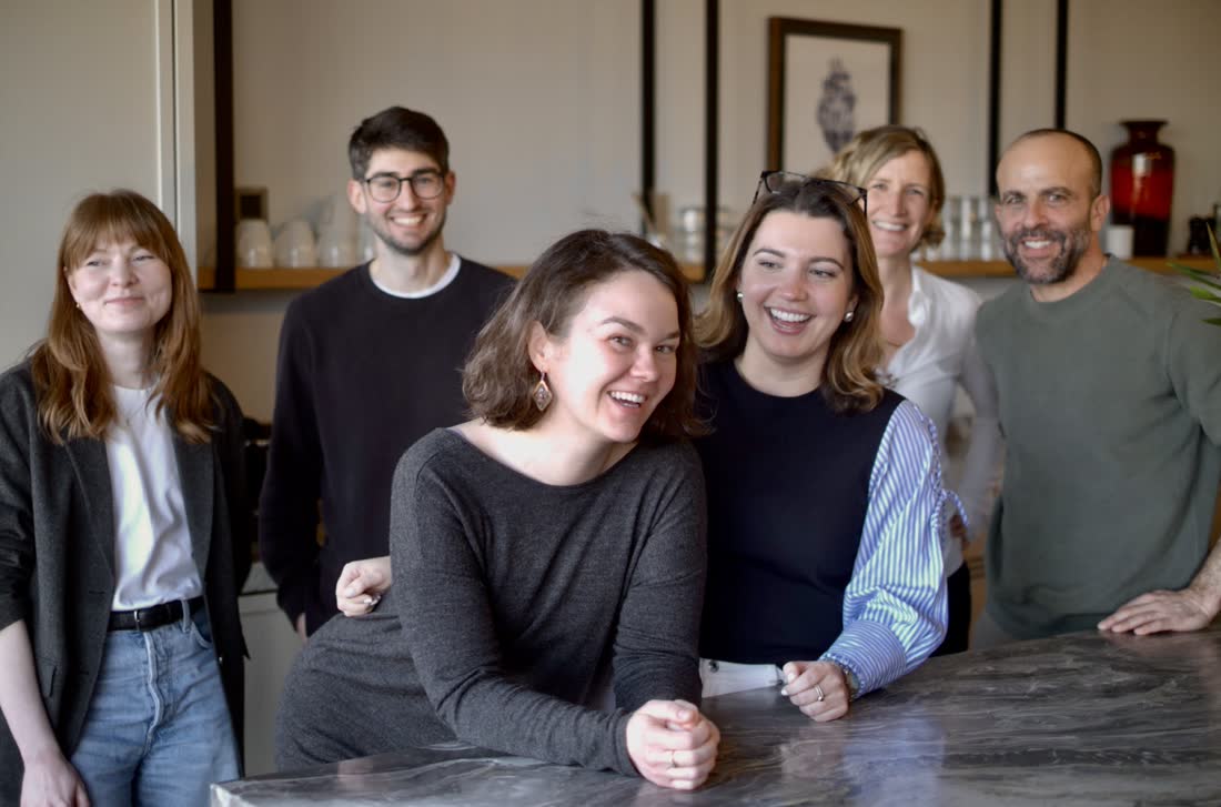 Six members of the team gathered and laughing around the marble counter in the studio kitchen.