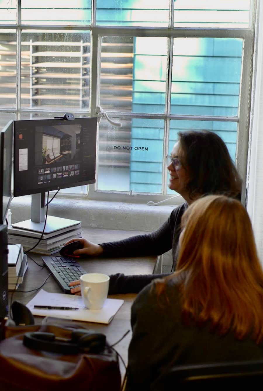 Two colleagues reviewing project photographs on screen at a desk by a large factory window.