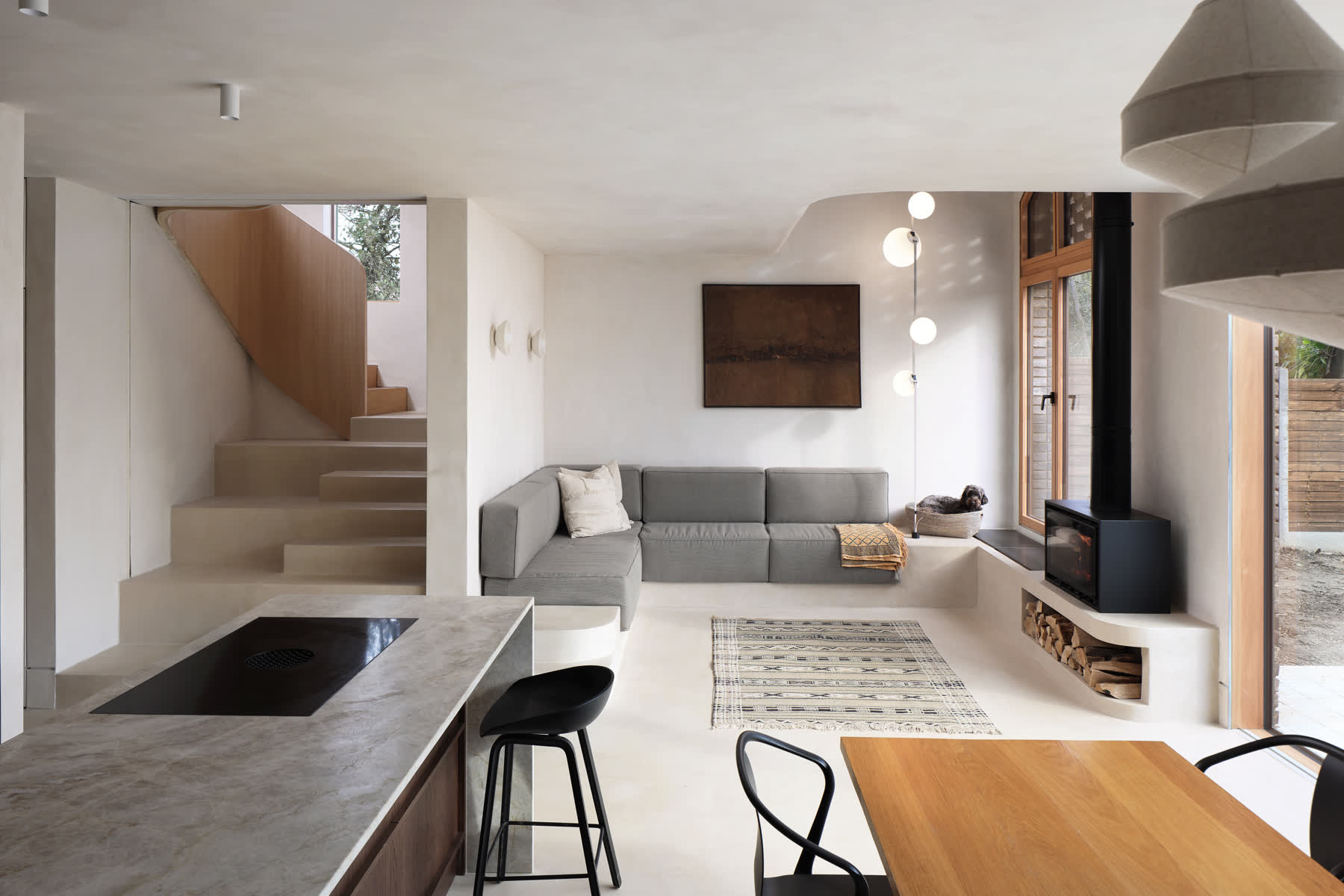 An open plan living, kitchen and dining space in pale plaster, with a cast stair and curved timber balustrade, a grey corner sofa and a wood burning stove beside garden doors