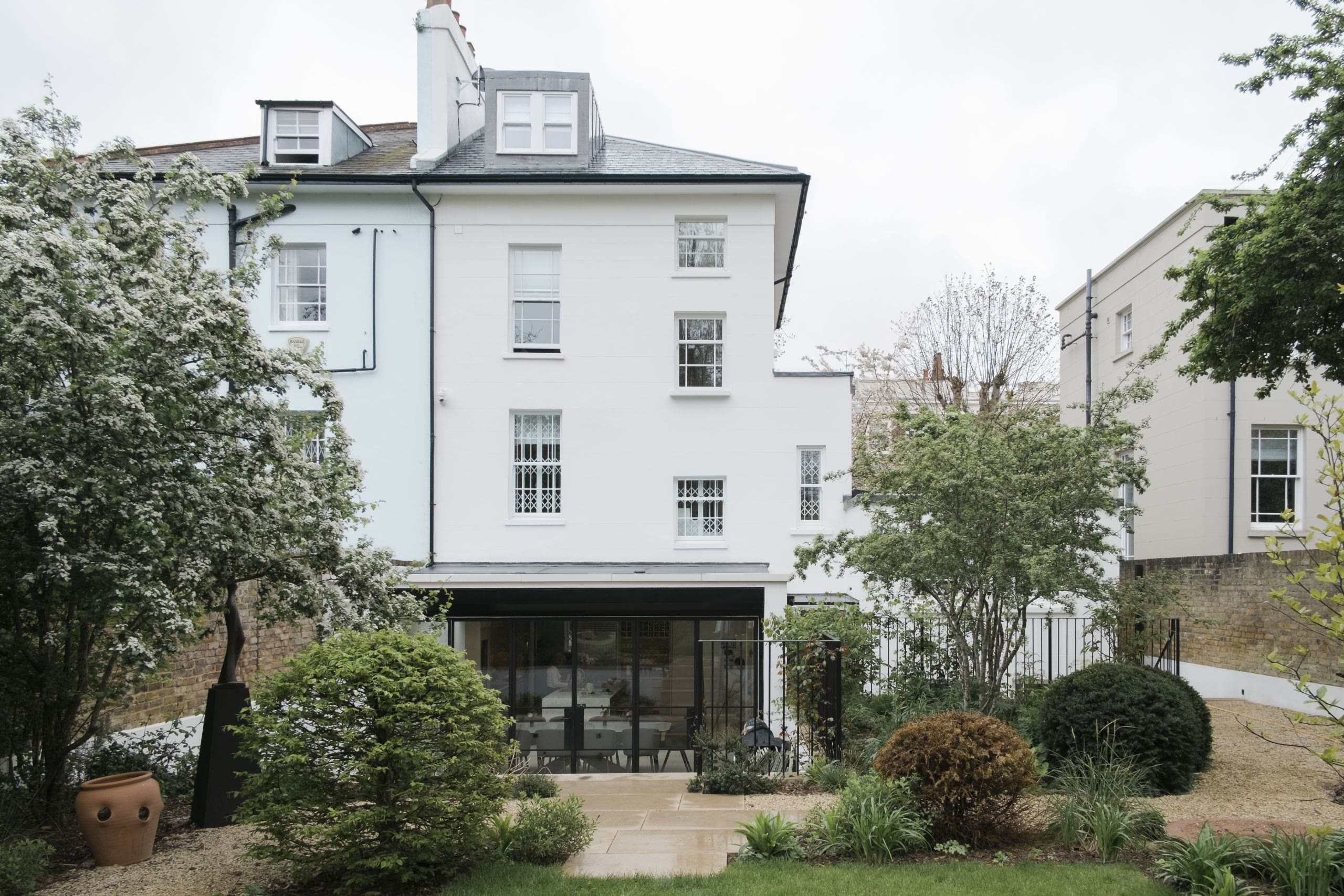The rear elevation of a white painted period house seen from the garden, with a dark framed glazed extension opening onto a stone path