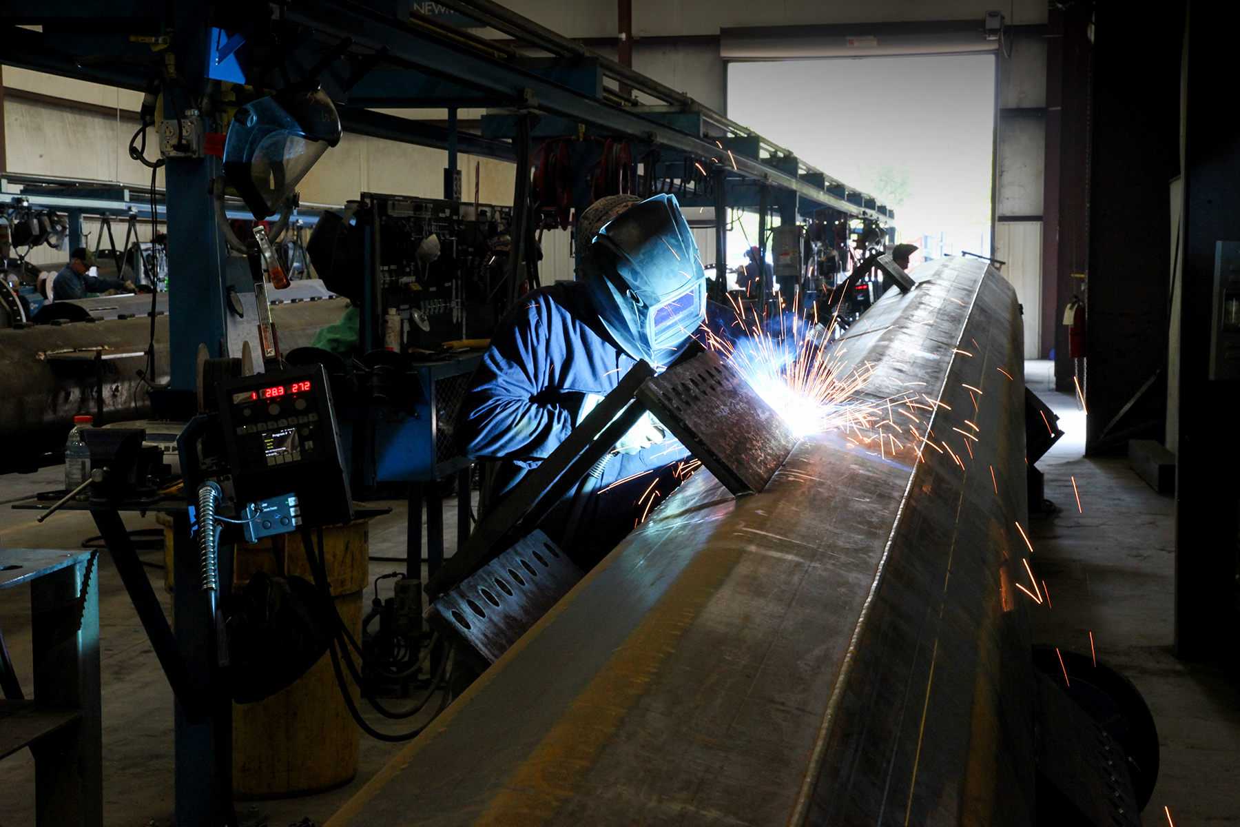 Worker in a welding helmet welds a large steel beam as sparks fly in a fabrication shop | Westside Construction Group