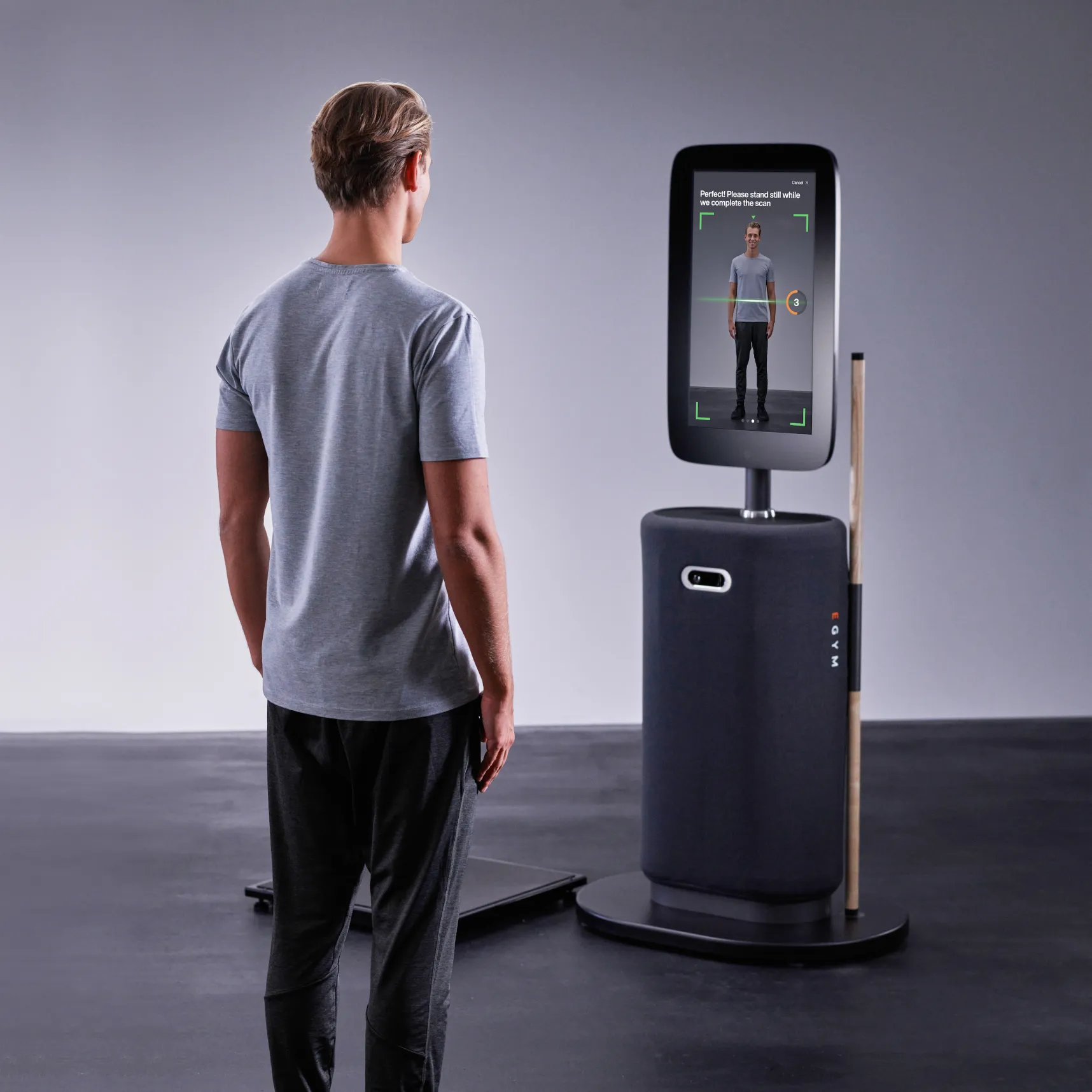 Man in gray t-shirt and black pants standing in front of a body scanning machine displaying a full-body scan with instructions to stand still.