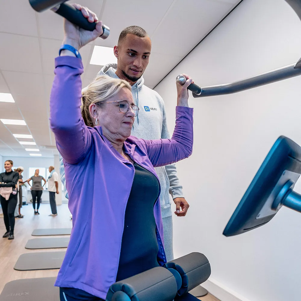 Senior woman in glasses exercising on a lat pulldown machine with a male trainer spotting her in a gym.