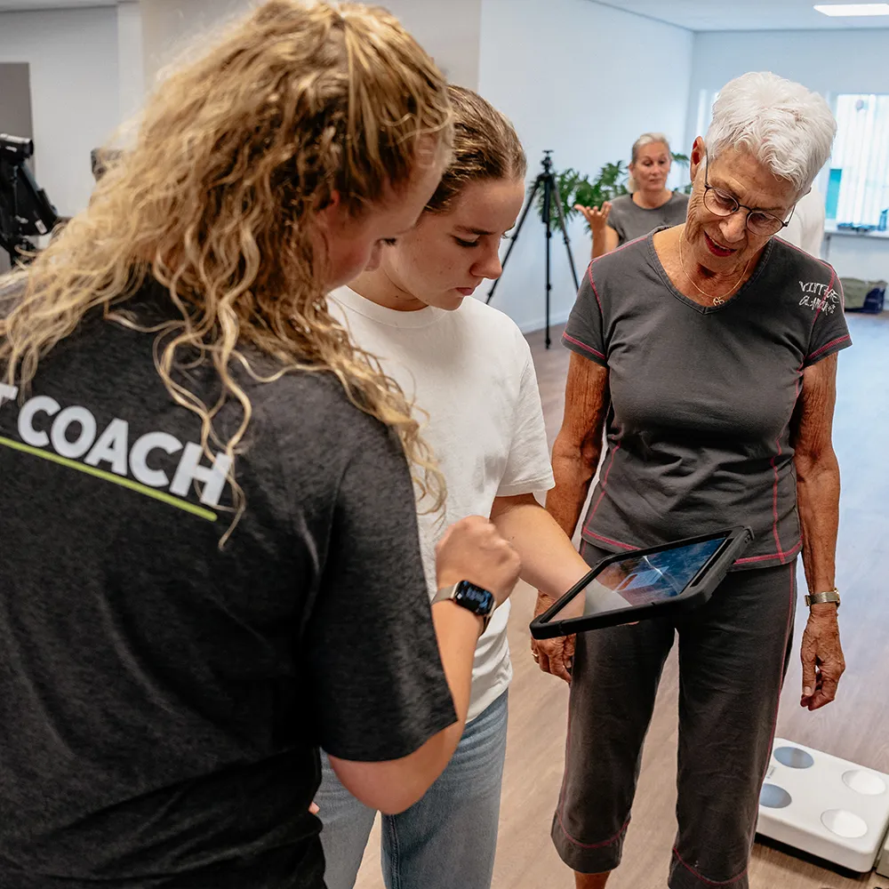 A fitness coach with curly hair shows a tablet to a young woman and an elderly woman in a workout room.