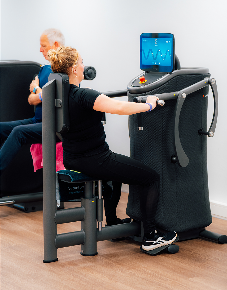 Woman using a seated rowing machine in a gym with a digital screen, while an older man sits on another exercise machine in the background.