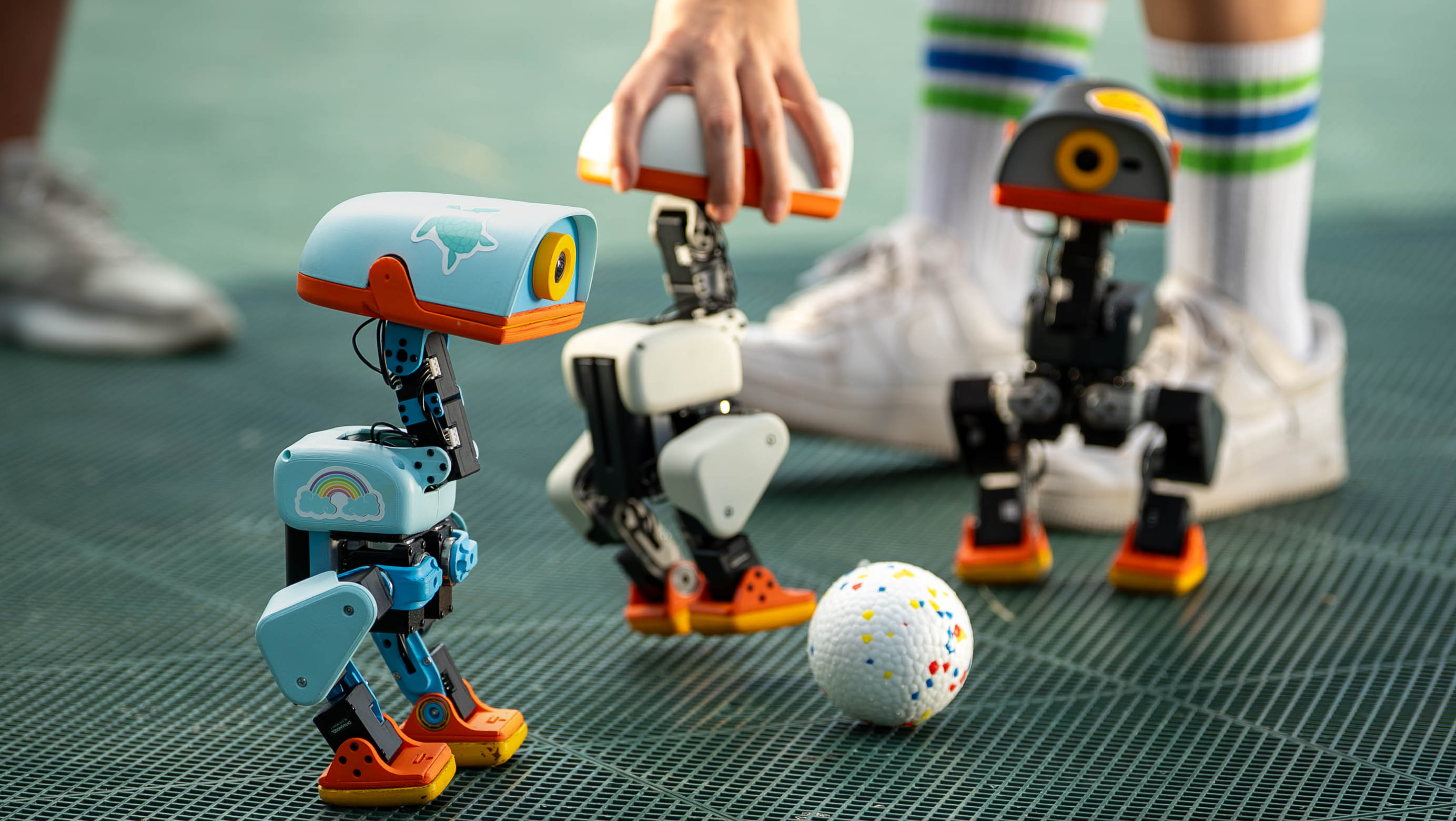 Three Microduck robots gathered around a ball, with a hand steadying the central robot.