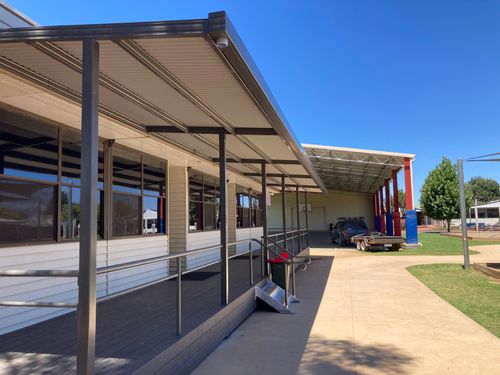 Covered walkway by a building with windows and a parked truck trailer under a metal shelter.
