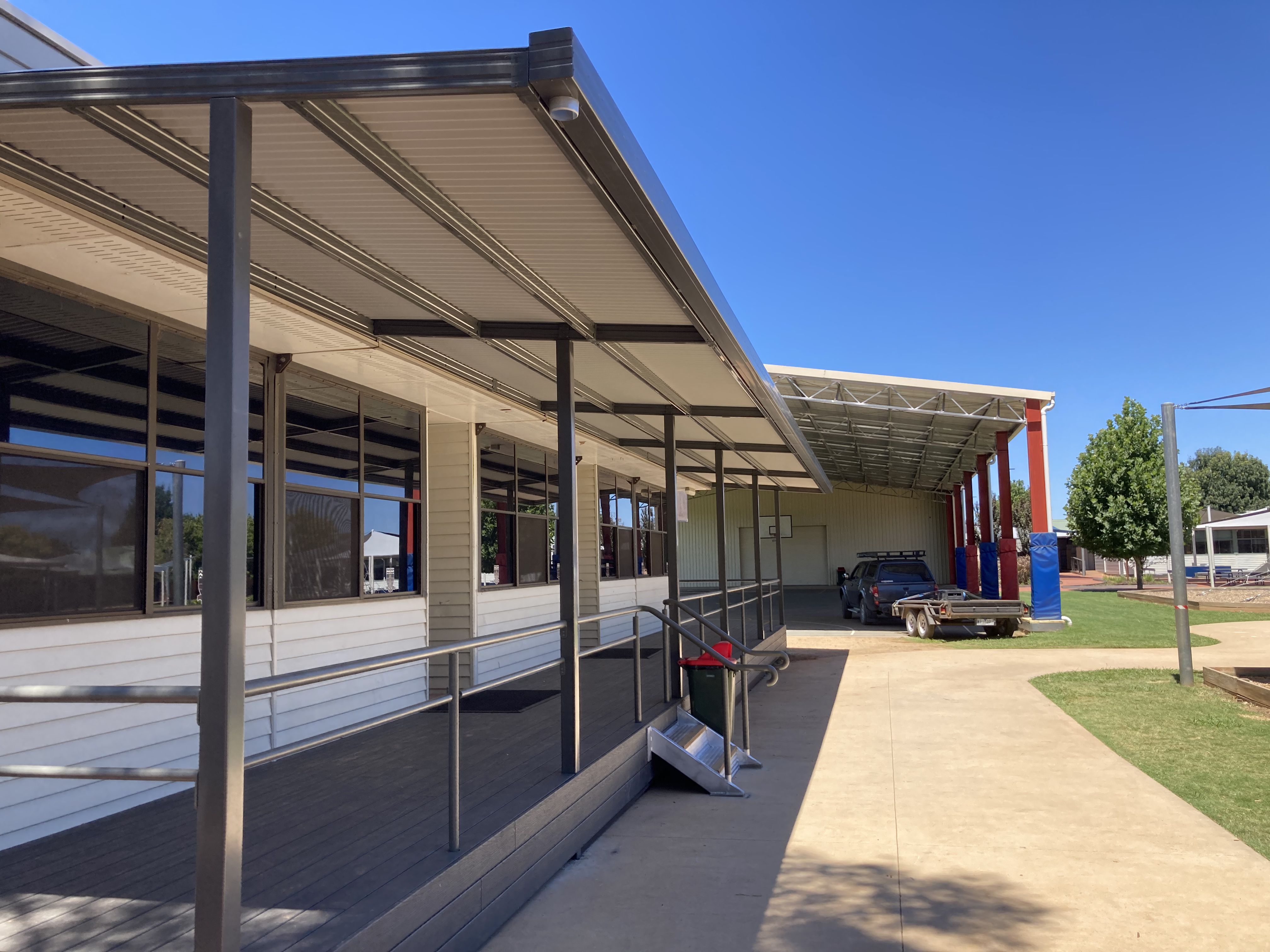 Covered walkway by a building with windows and a parked truck trailer under a metal shelter.