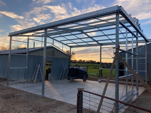 Metal frame structure being built next to metal sheds on concrete floor at sunset with green field background.