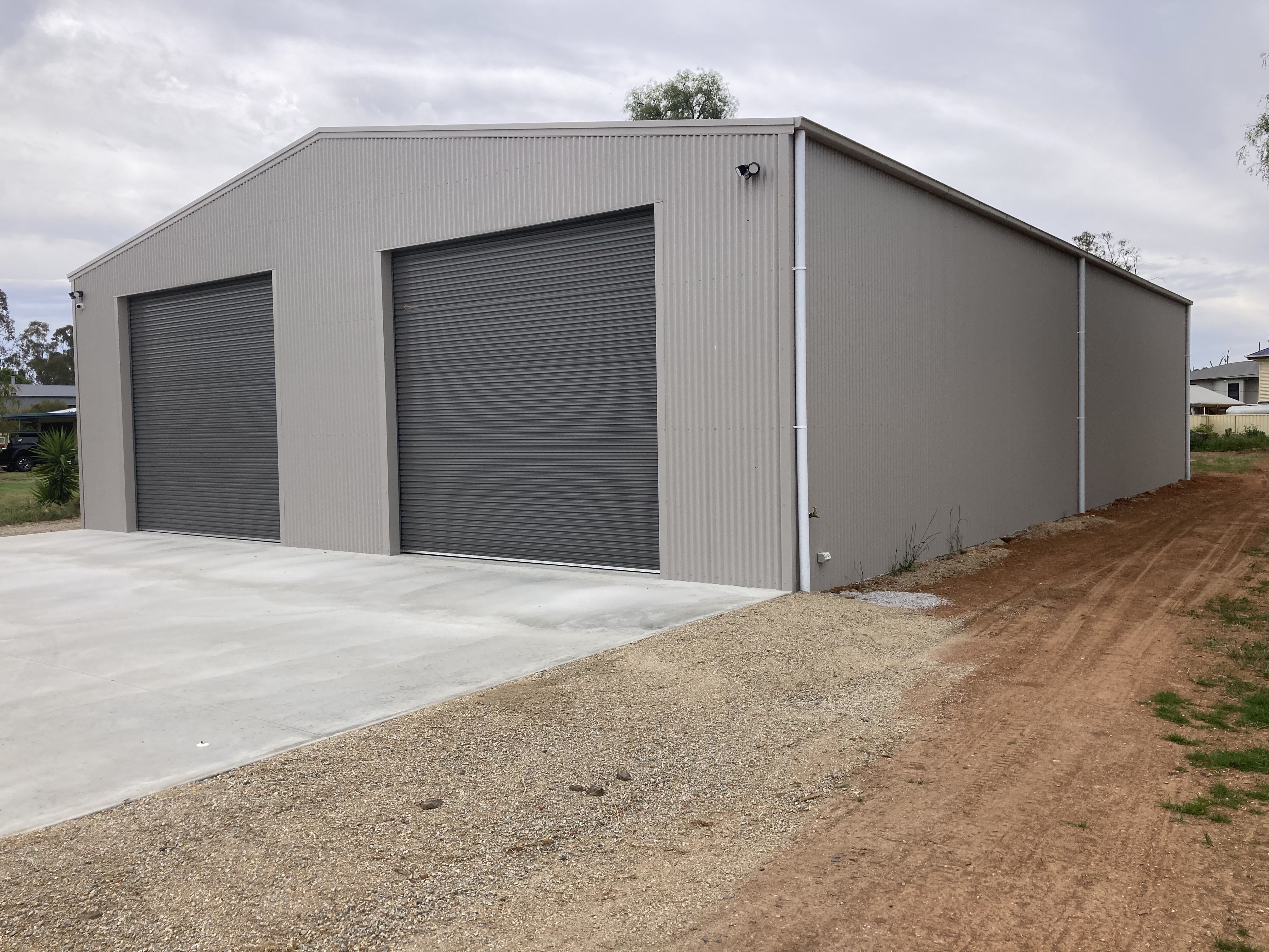 Large light gray metal building with two dark gray closed roll-up doors and gravel dirt driveway.